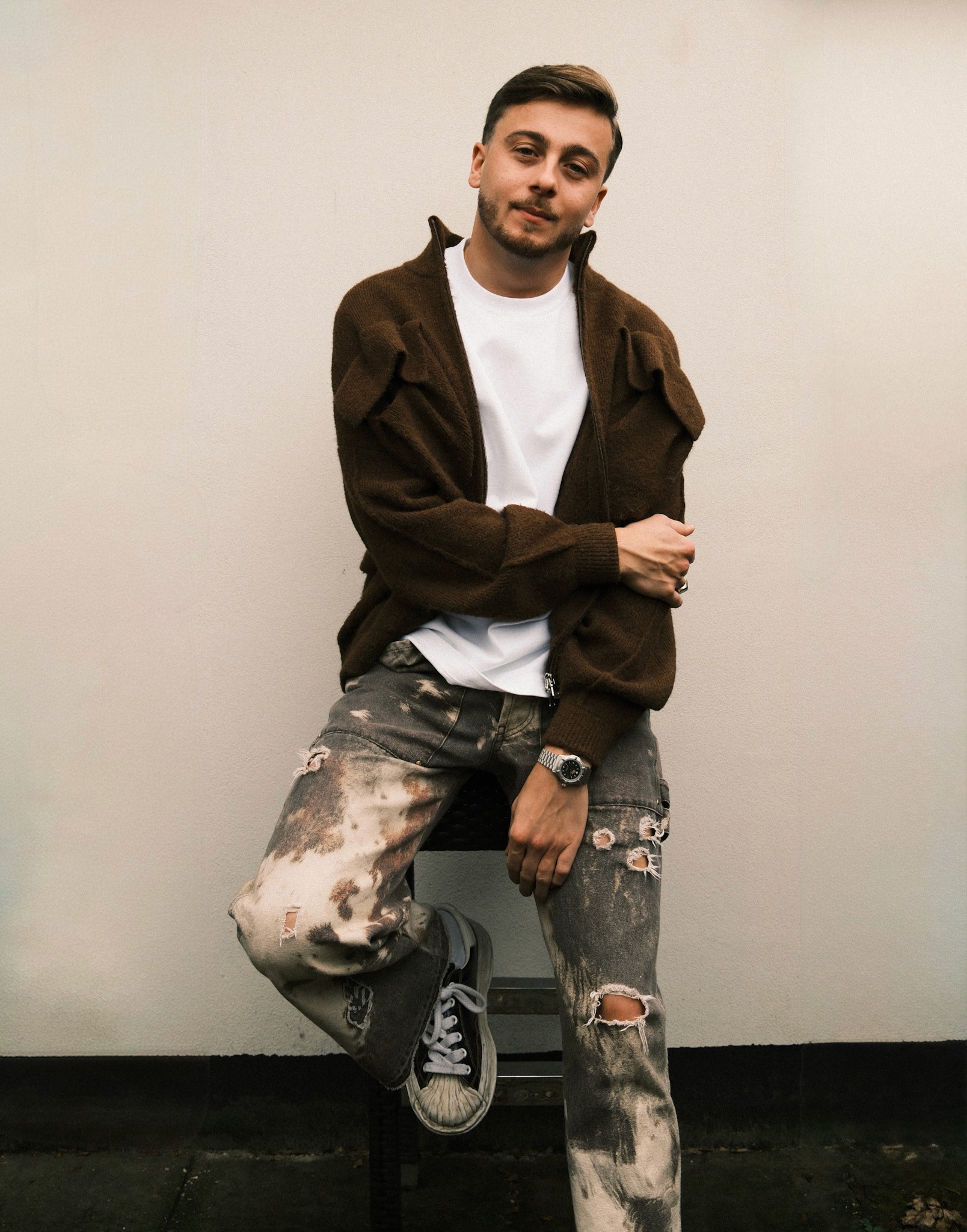 Portrait en pied de Sem Jacobs posant devant un mur blanc, portant un blouson marron ouvert sur un t-shirt blanc, un jean délavé avec des déchirures et des baskets sombres.