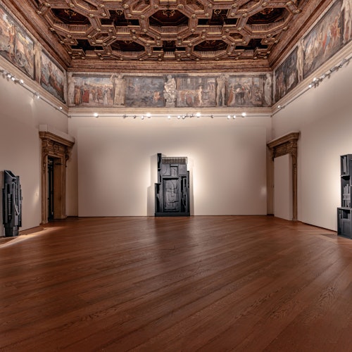 Una galleria d'arte con pavimenti in legno, soffitto decorato, affreschi alle pareti e sculture astratte nere su pareti bianche.