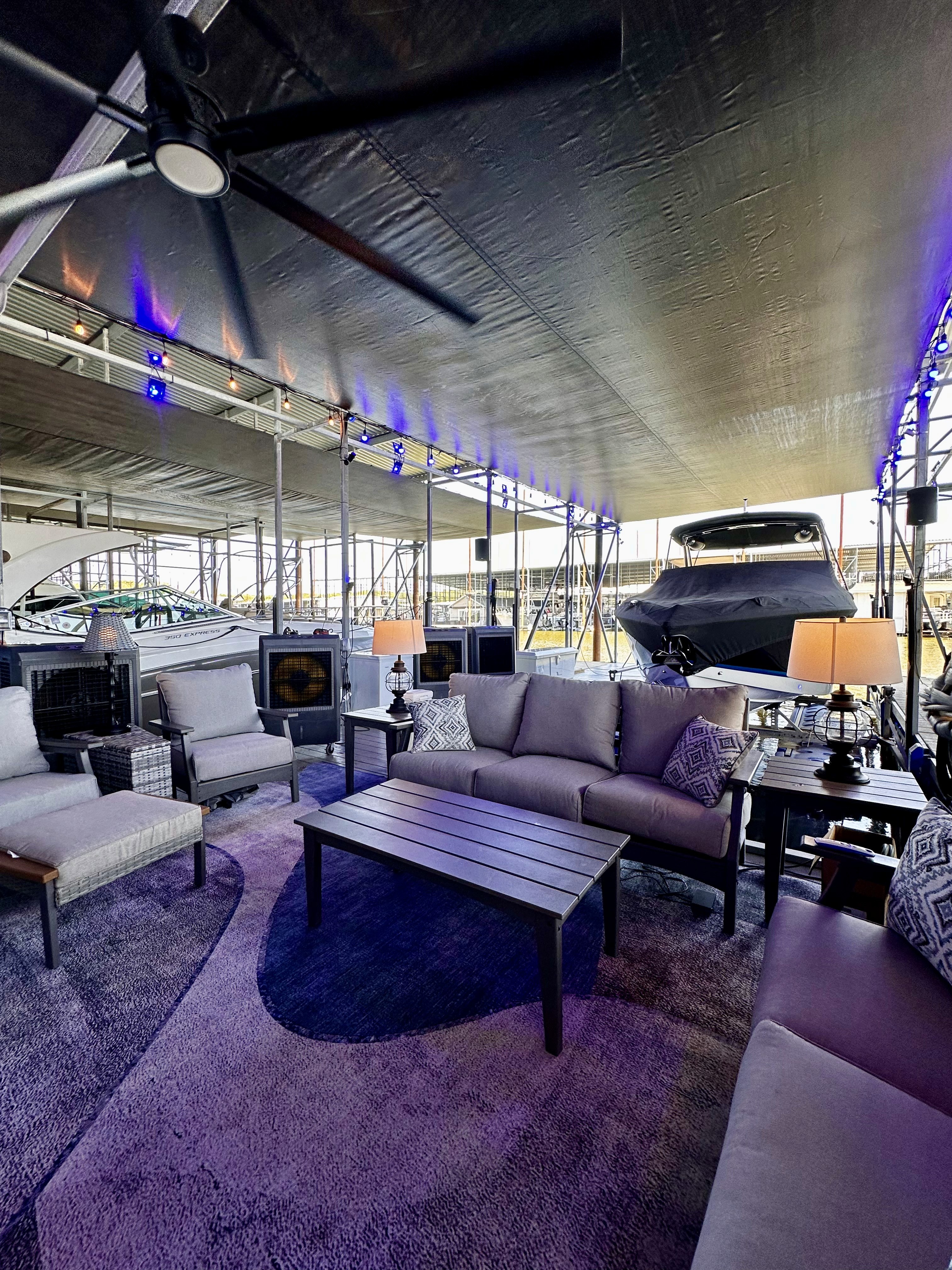 A modern, covered marina lounge area features plush outdoor seating, a long wooden coffee table, ambient lighting, and boats docked in the background, creating a luxurious and relaxing waterfront atmosphere.