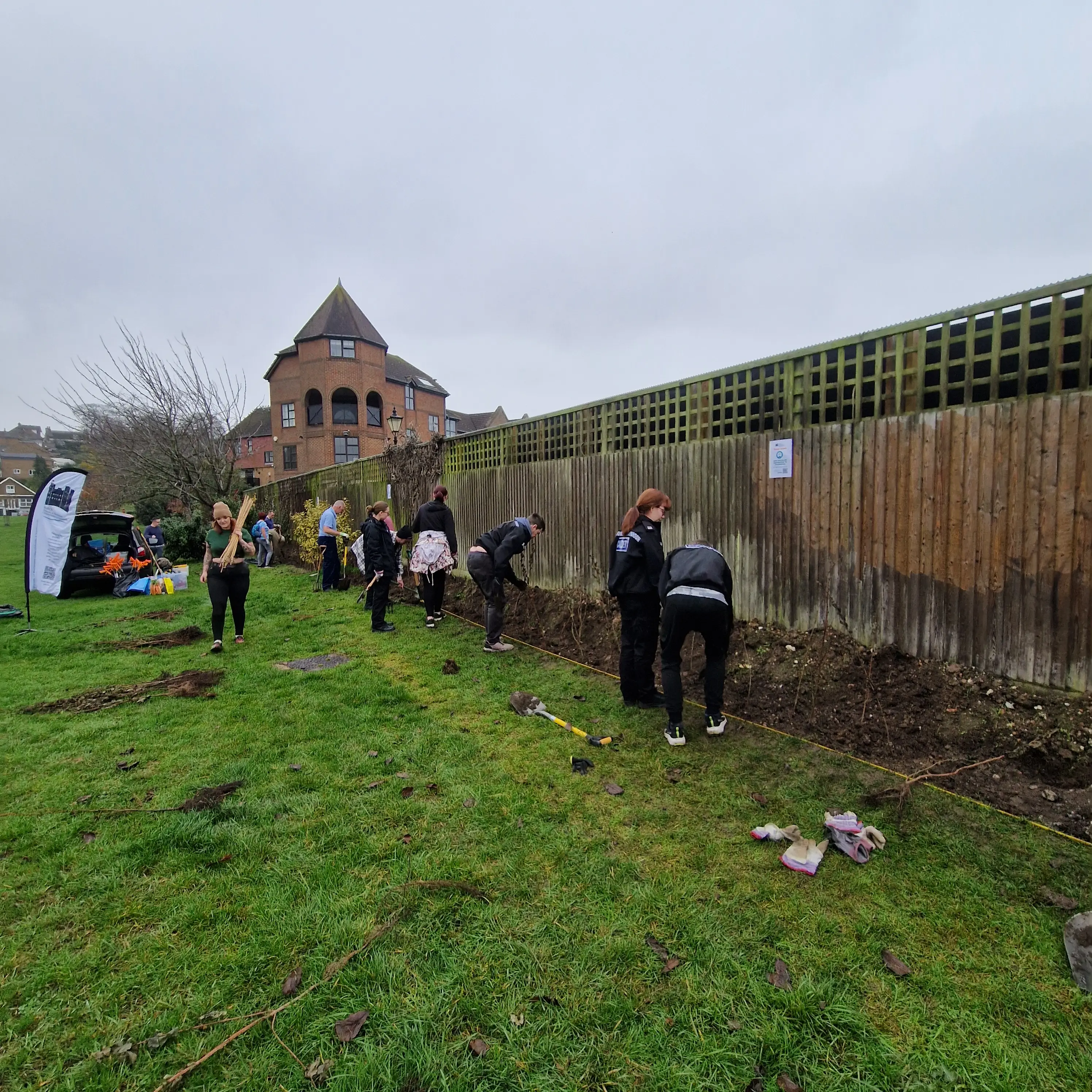 People working together along a fence in a grassy area, with a building visible in the background.