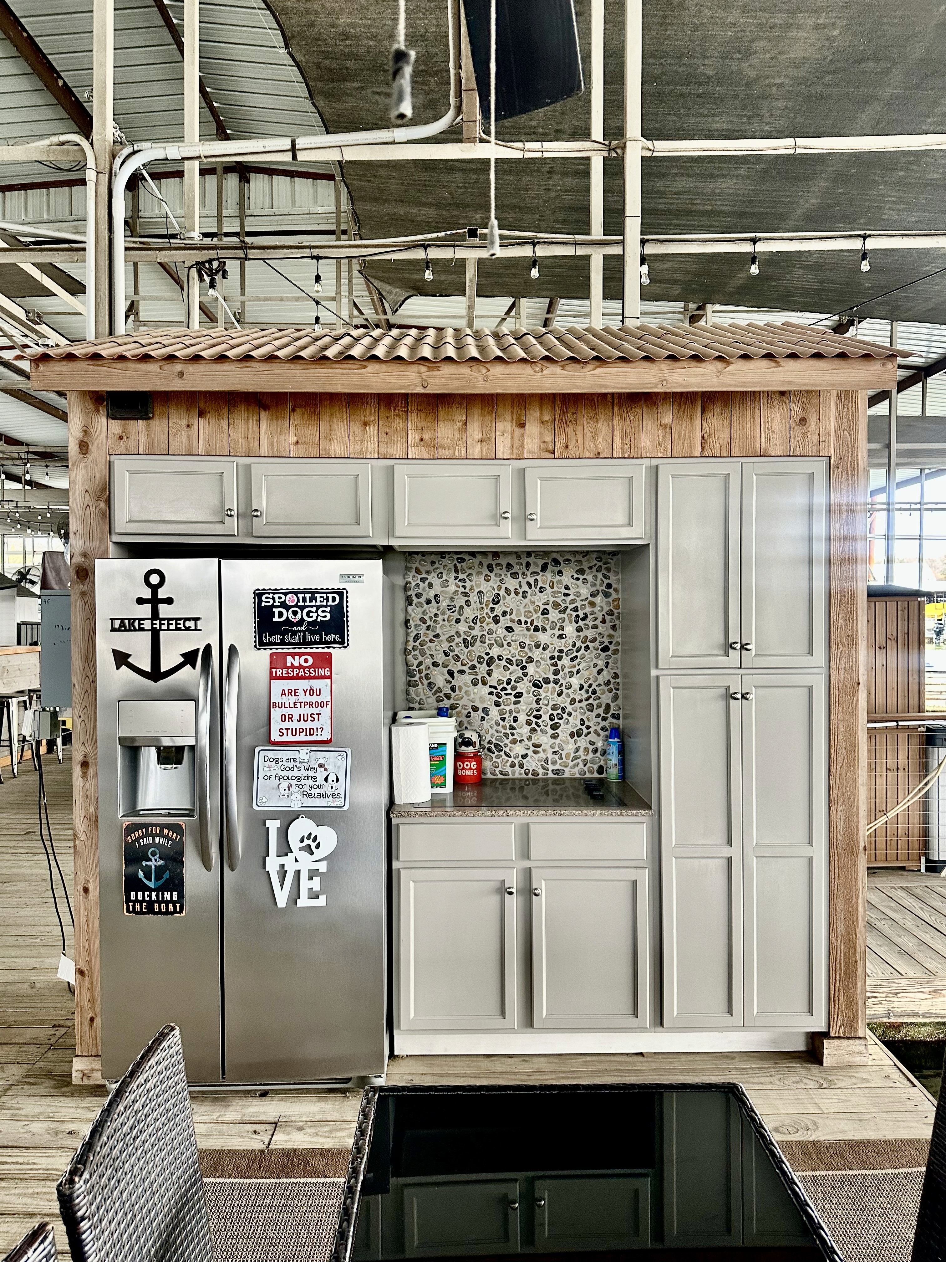A modern outdoor kitchenette under a large, industrial roof features sleek gray cabinets, a stainless steel refrigerator with playful magnets and a sign prohibiting dogs, a small mosaic backsplash, and bottles of dish soap placed by the sink.