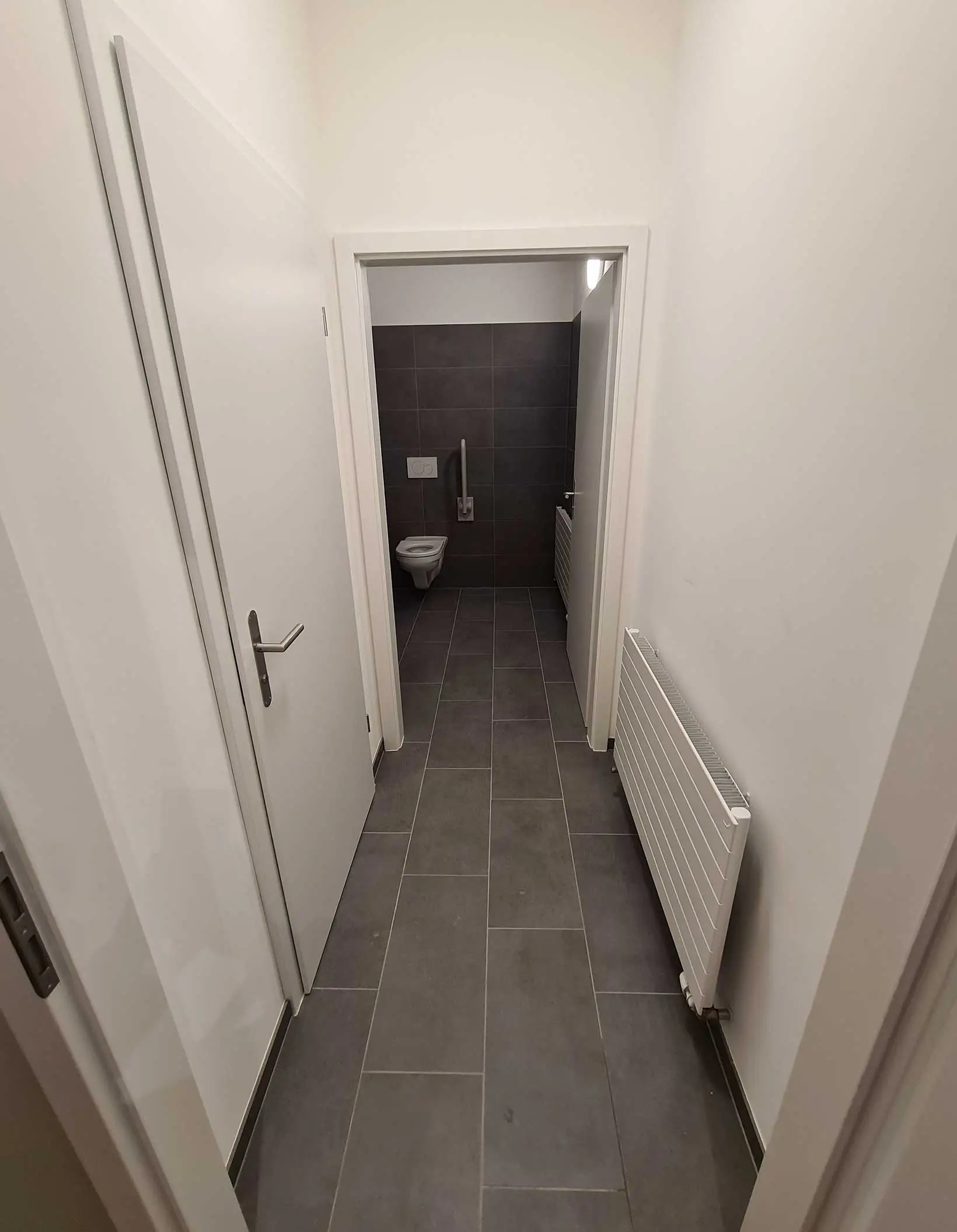 Couloir menant à des toilettes entièrement carrelée de gris foncé, avec une porte ouverte donnant sur un toilette.