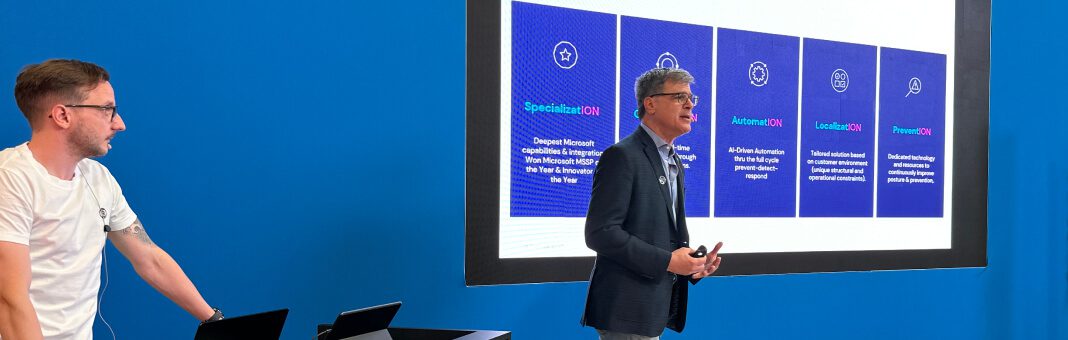 Zwei Vortragende in einer professionellen Umgebung, wobei ein Mann spricht und der andere unterstützt. Auf einem großen Bildschirm werden fünf blau hervorgehobene Abschnitte mit Informationen zu einem Geschäftsthema angezeigt.