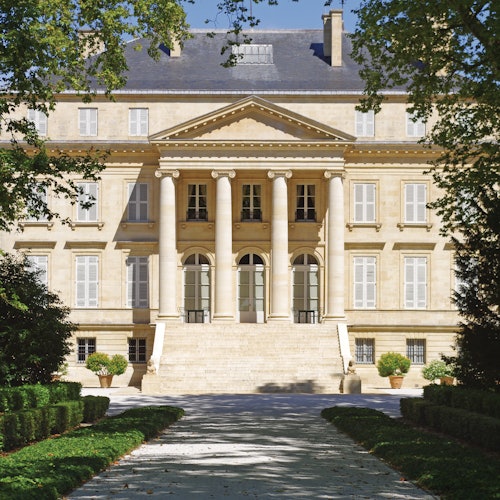 Grand neoclassical building with six columns, arched windows, and wide stone steps. Framed by manicured hedges and trees.