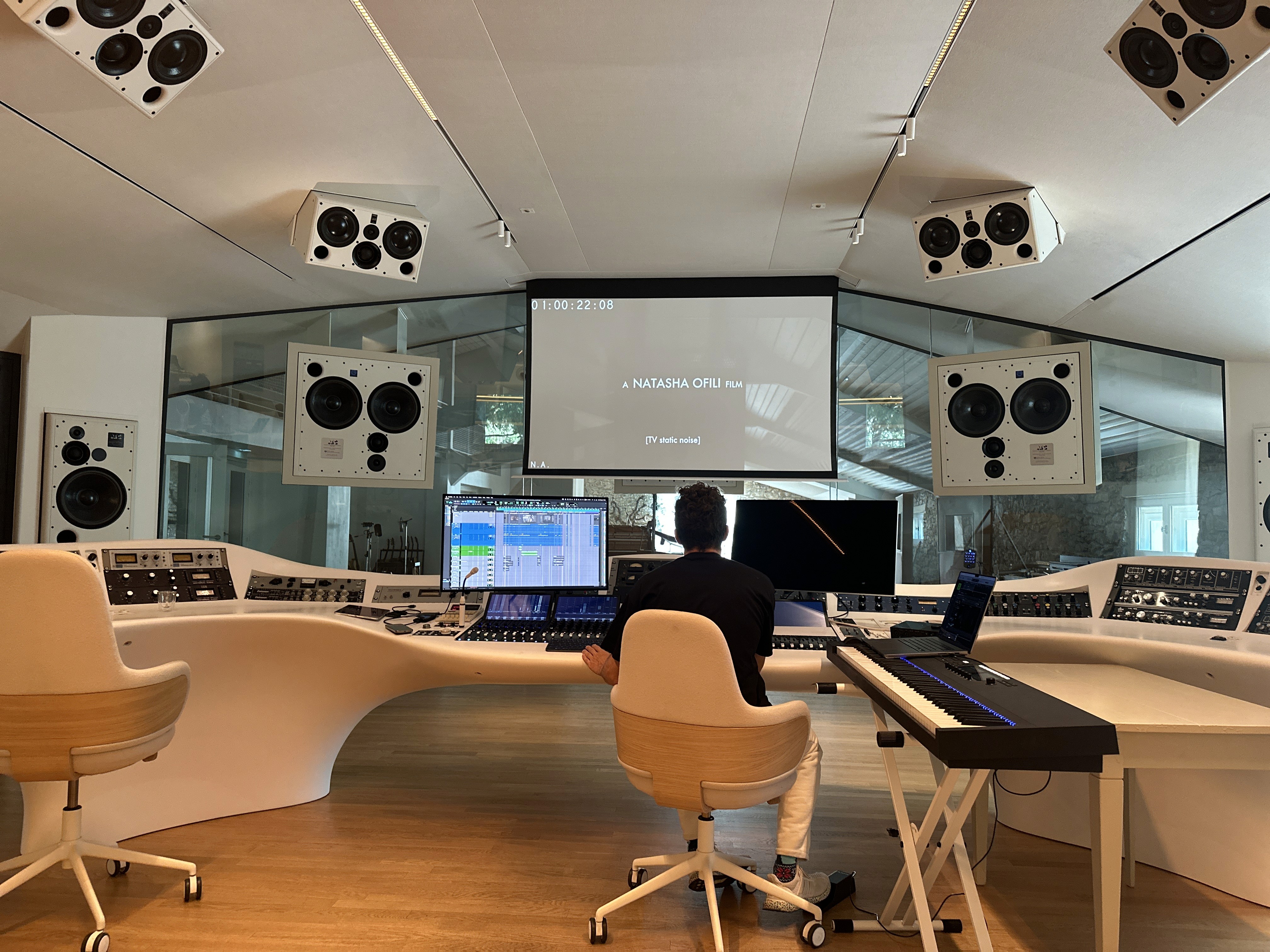 Damien Quintard in Miraval Studios’s modern recording studio with a large control desk, speakers, and a monitor displaying audio software. A projector screen shows text: A Natasha Ofili Film (TV static noise).