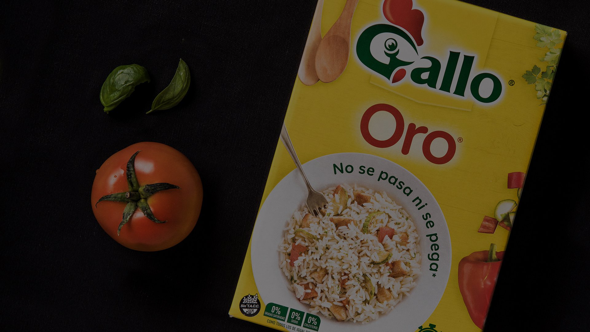Yellow box of Gallo Oro rice with a fork and rice image. Tomato and basil leaves are on the side on a dark surface, conveying a fresh, natural feel.