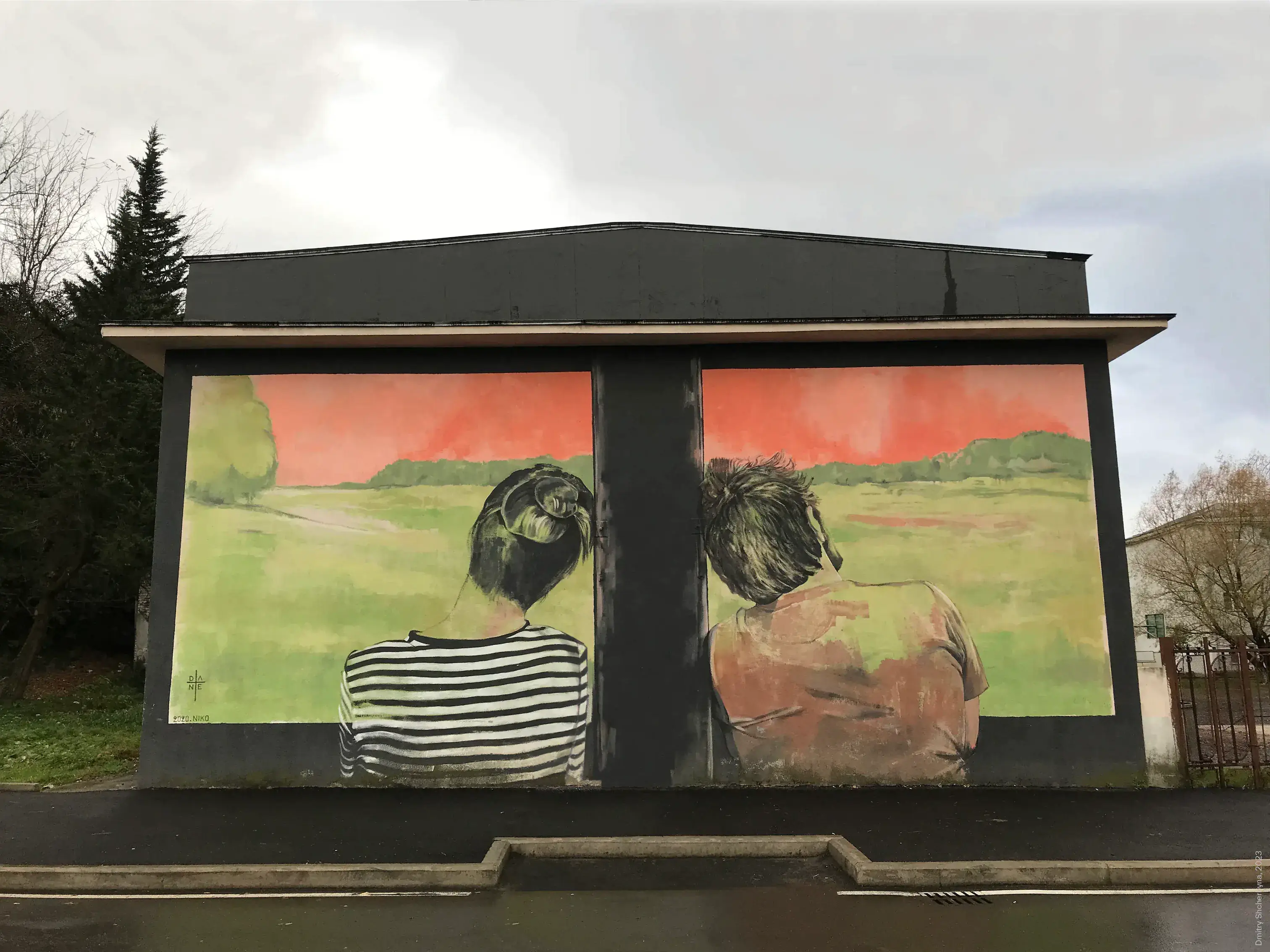 A mural painted on a small building shows the backs of a woman and a man gazing out at a green field. Their views are separated so each see their own picture.