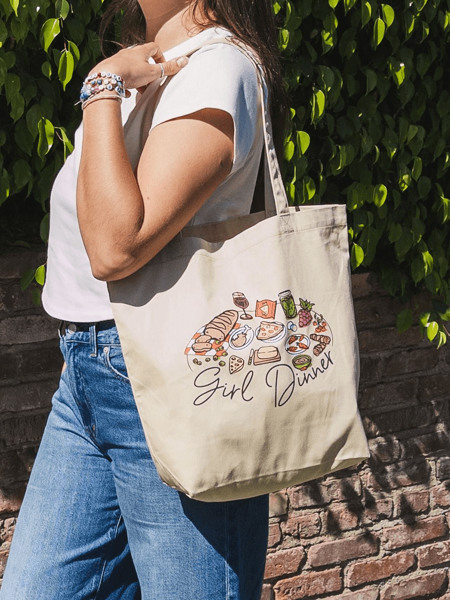 Woman holding tote bag with "Girl Dinner" design, casual outfit, outdoor background.