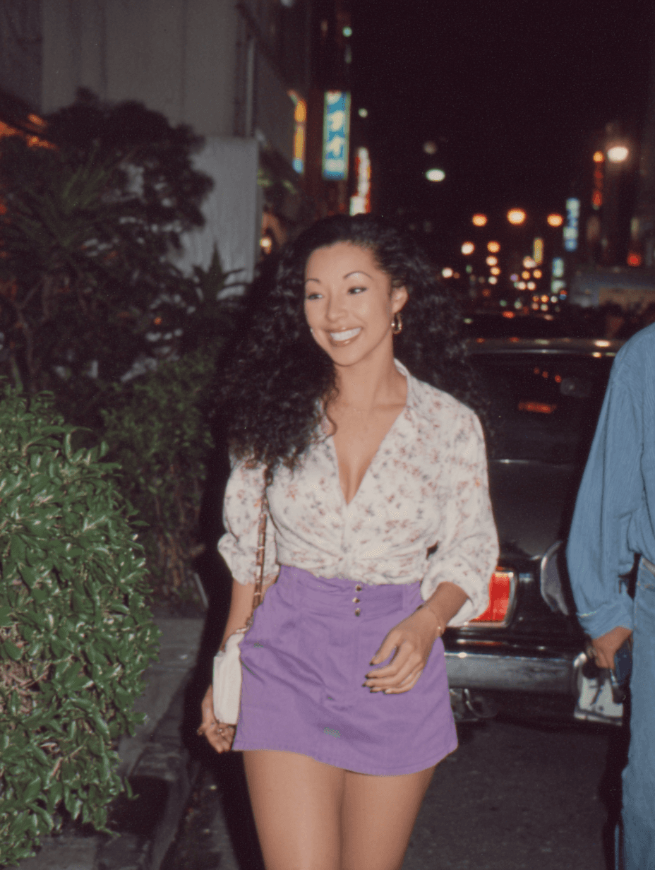 Stylish woman with voluminous curly hair walking through a neon-lit Tokyo street at night, wearing a floral blouse and purple skirt with a confident smile.