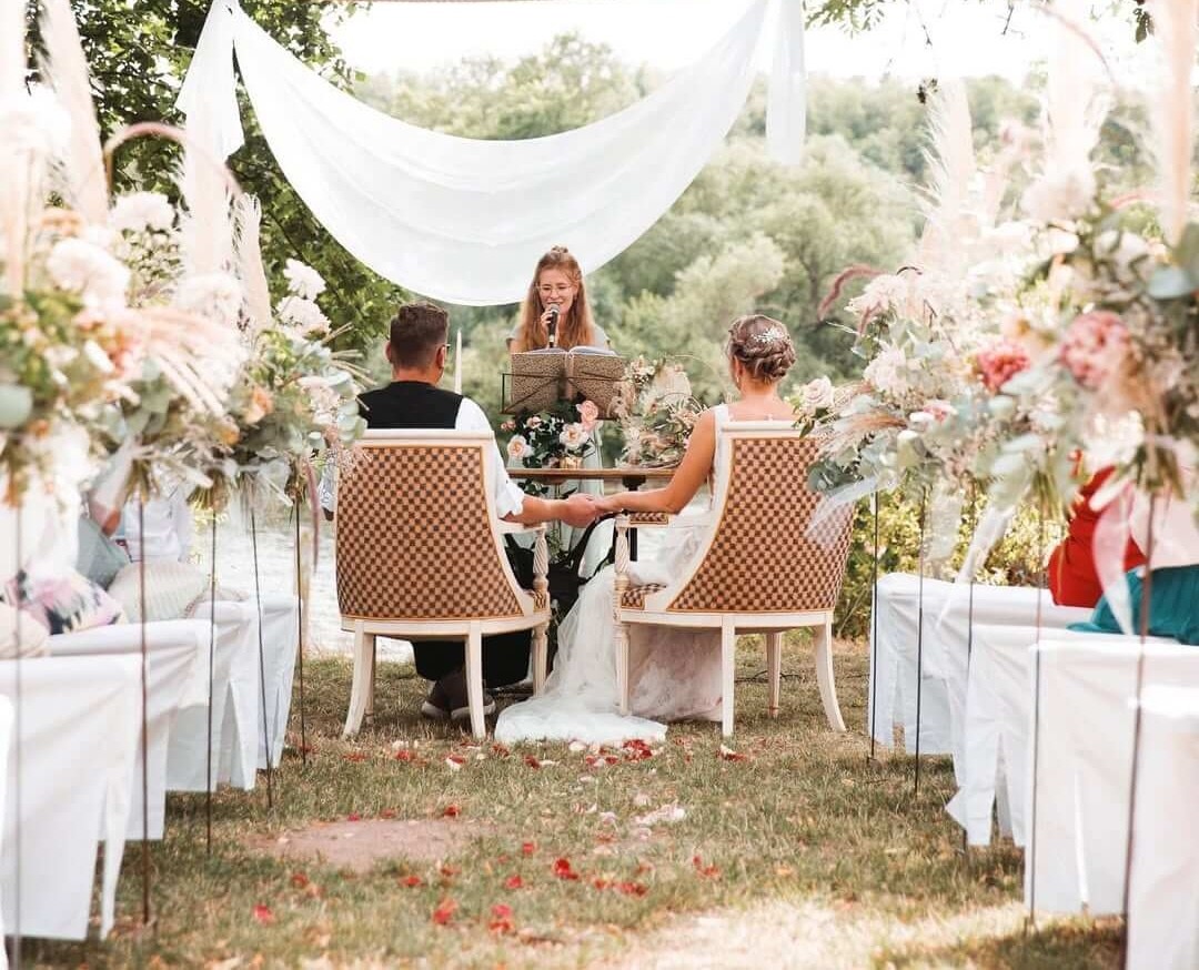 Brautpaar bei Hochzeitszeremonie unter goldenem Bogen, umgeben von Blumen. Frau liest, Braut trägt Schleier, Bräutigam im Anzug.