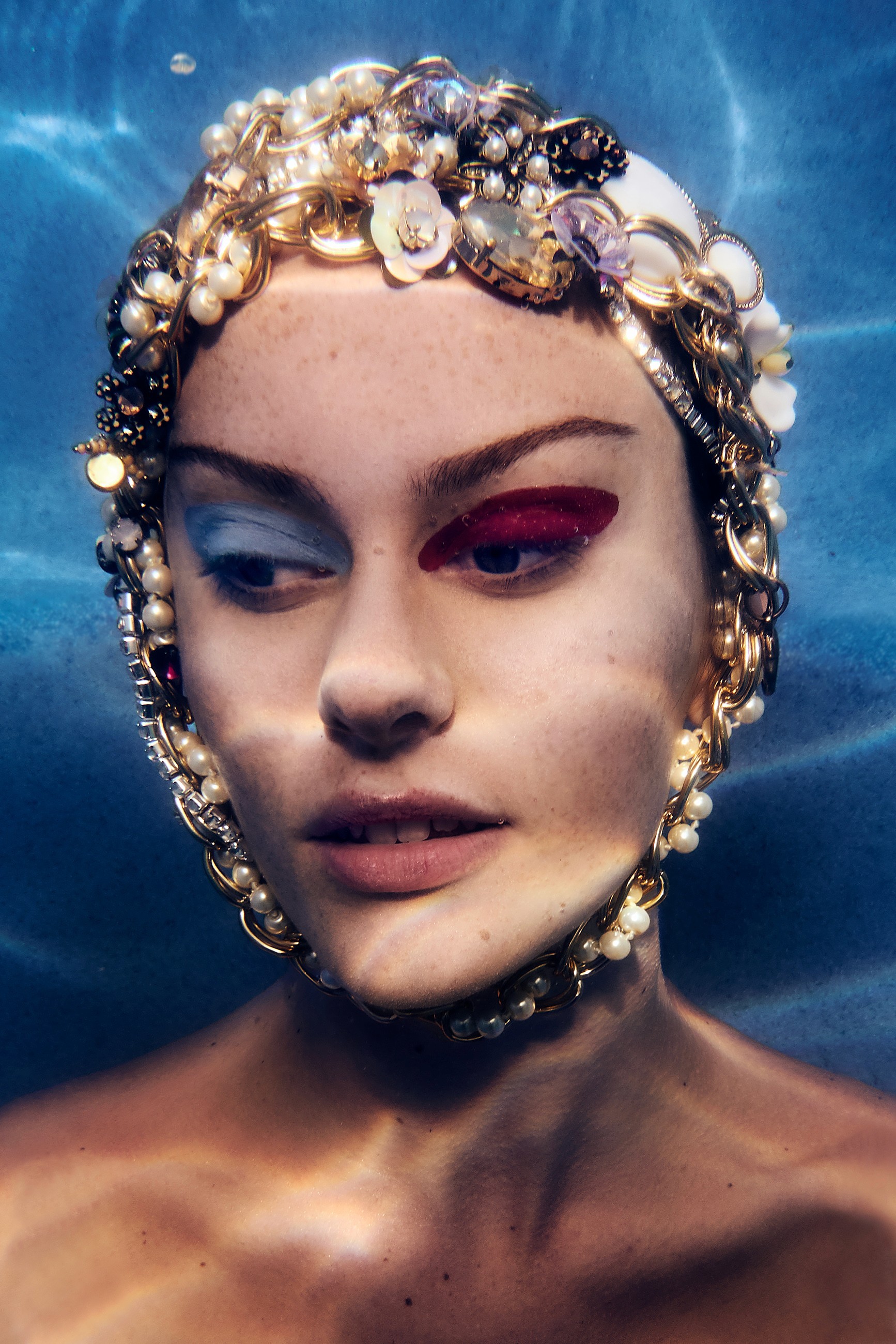 Underwater beauty photography of a model with her eyes open. The eye shadow makeup is blue on one eye and red on the other. She is wearing a swimming cap completely covered in sparkling jewels.