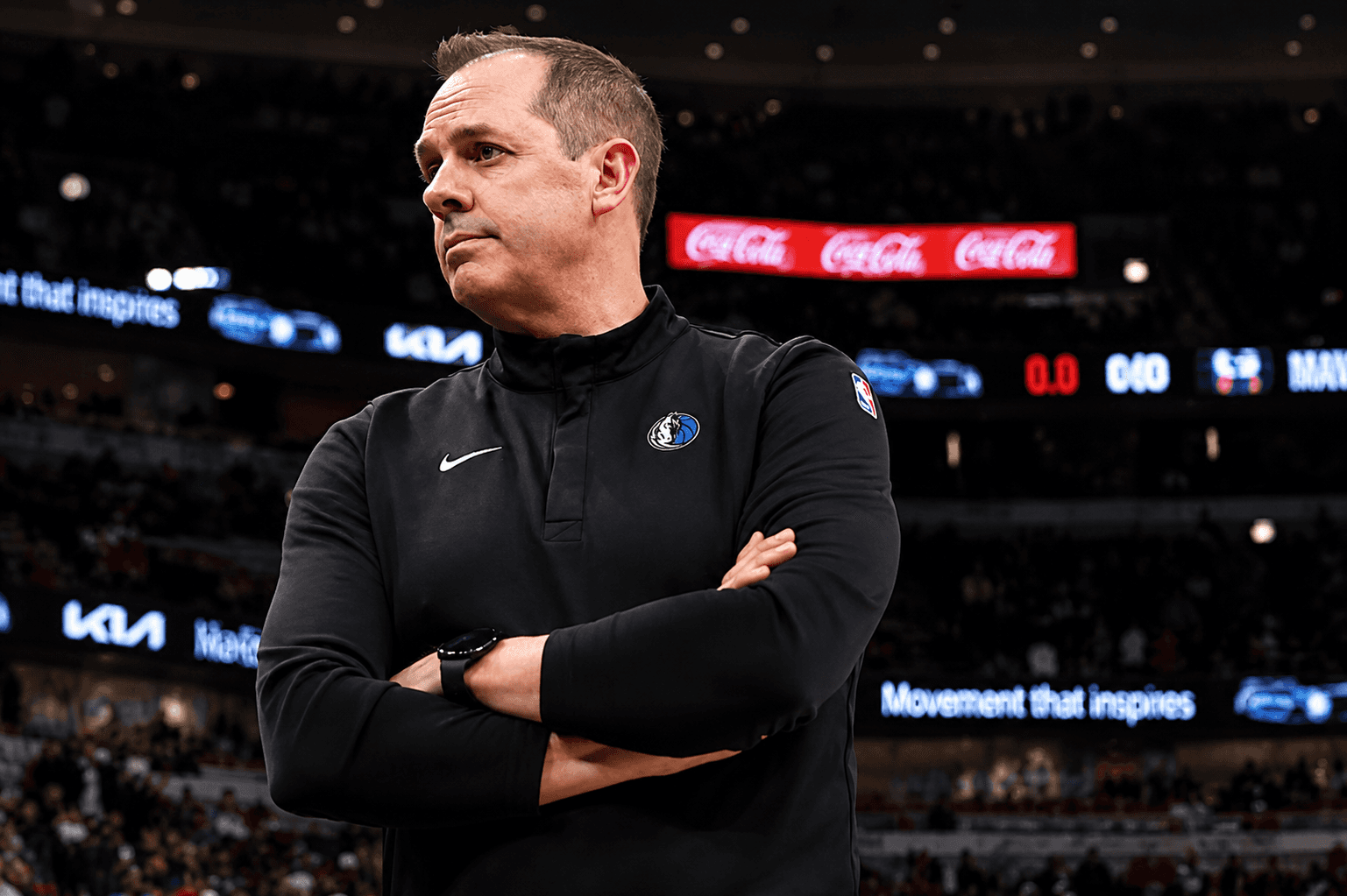 Frank Vogel wearing a black Dallas Mavericks pullover stands with arms crossed in an arena, with blurred spectators and electronic advertisements in the background.