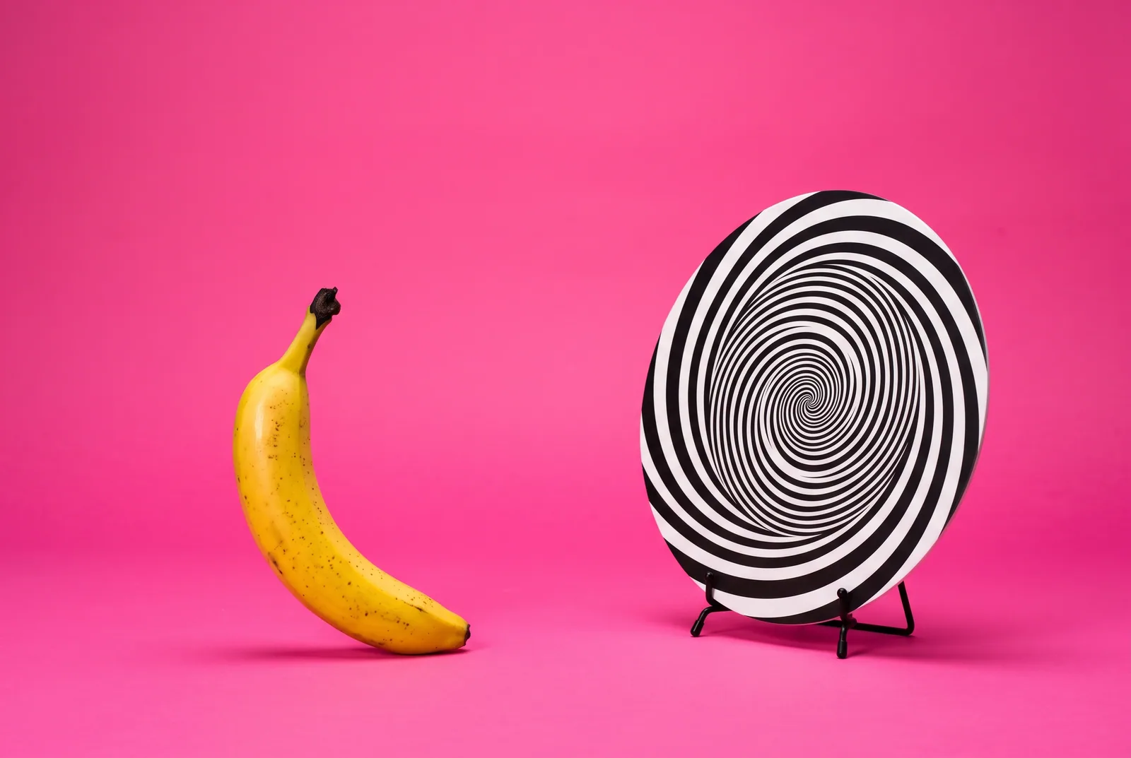 Gooning addiction concept: a ripe banana leaning against a large hourglass filled with pink sprinkles on a lilac backdrop
