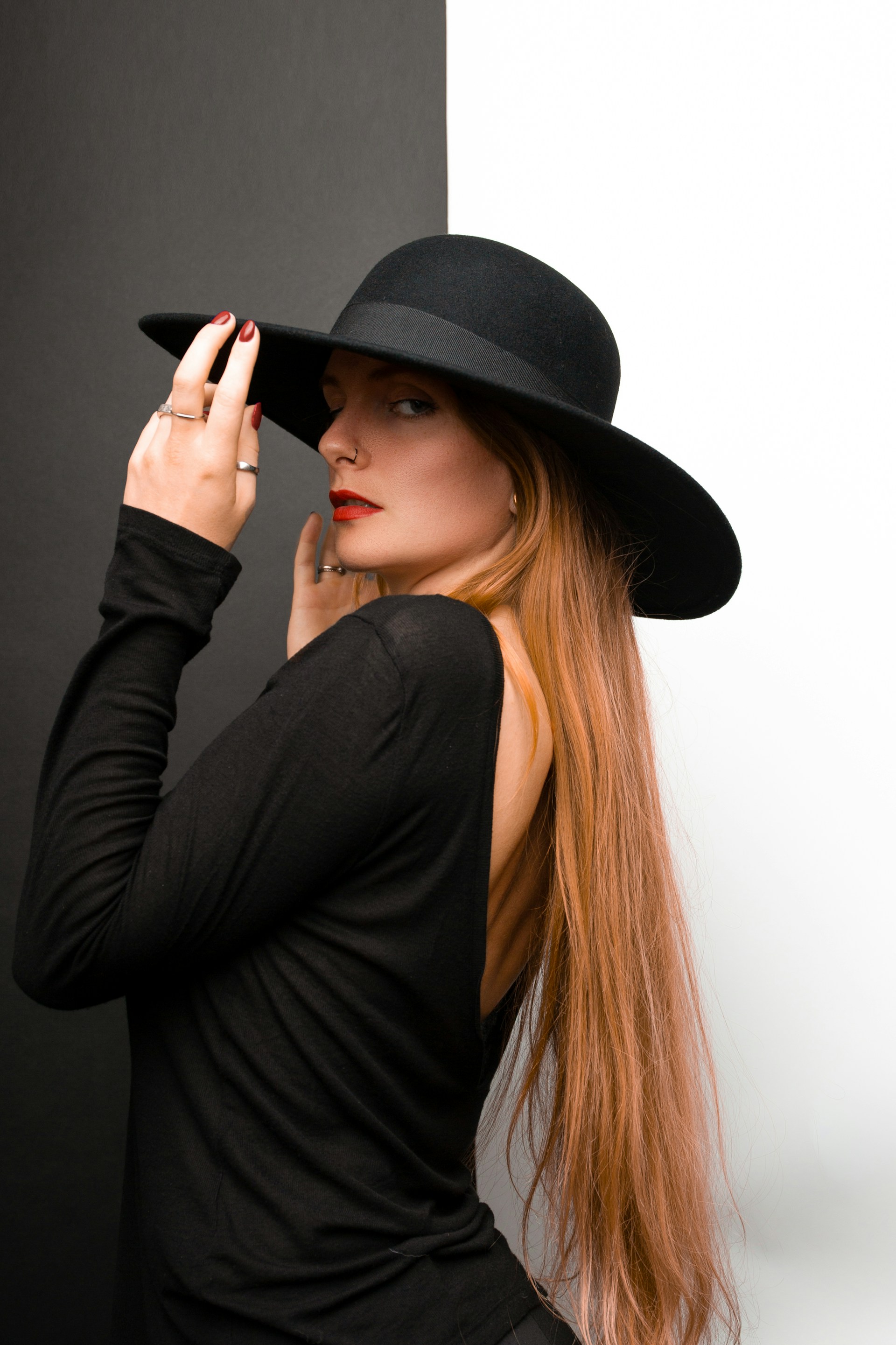 A side profile of a woman with long strawberry-blonde hair looking over her shoulder. She is wearing a black wide-brimmed hat, a backless black top, and bright red lipstick.