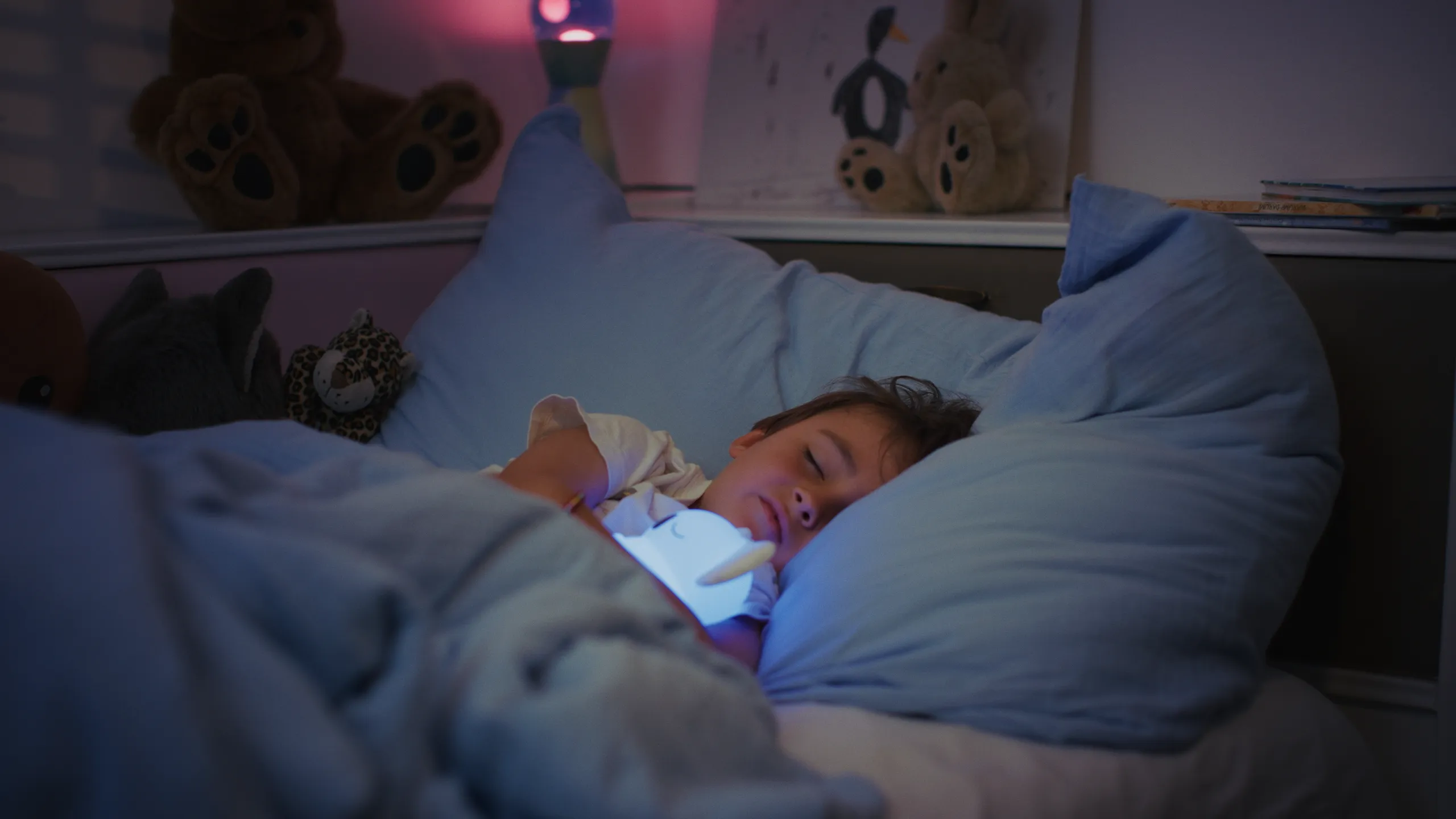 A peaceful shot of a young child sleeping in bed illuminated by a soft blue glow from a tablet or light source.