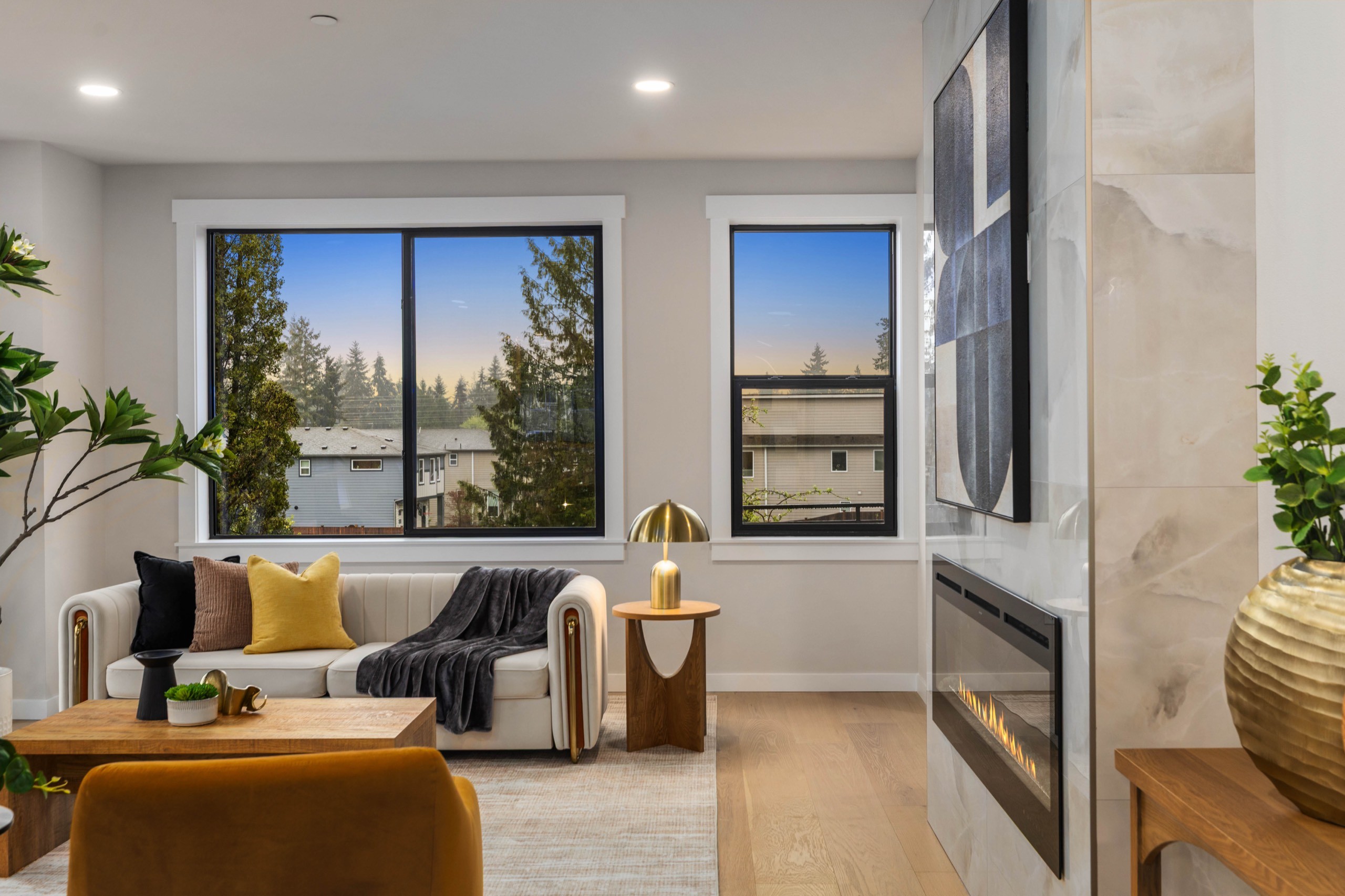 Serene luxury living room with panoramic lake and tree views, linear fireplace with stone surround, neutral linen sofa, amber accent chair, and wall-mounted TV
