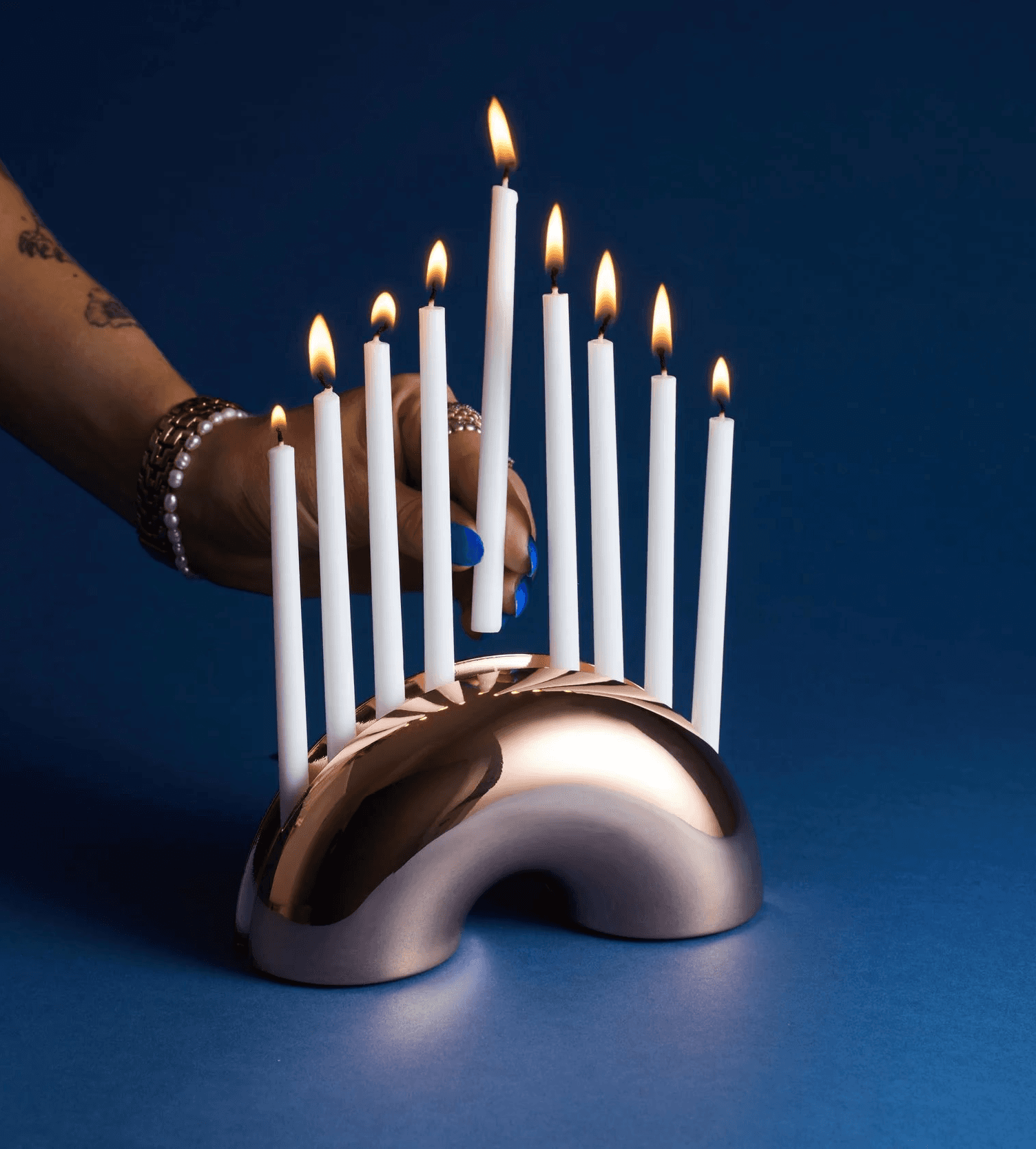 Hand reaching toward the lit Modern Mensch Nosh Menorah on a dark blue background