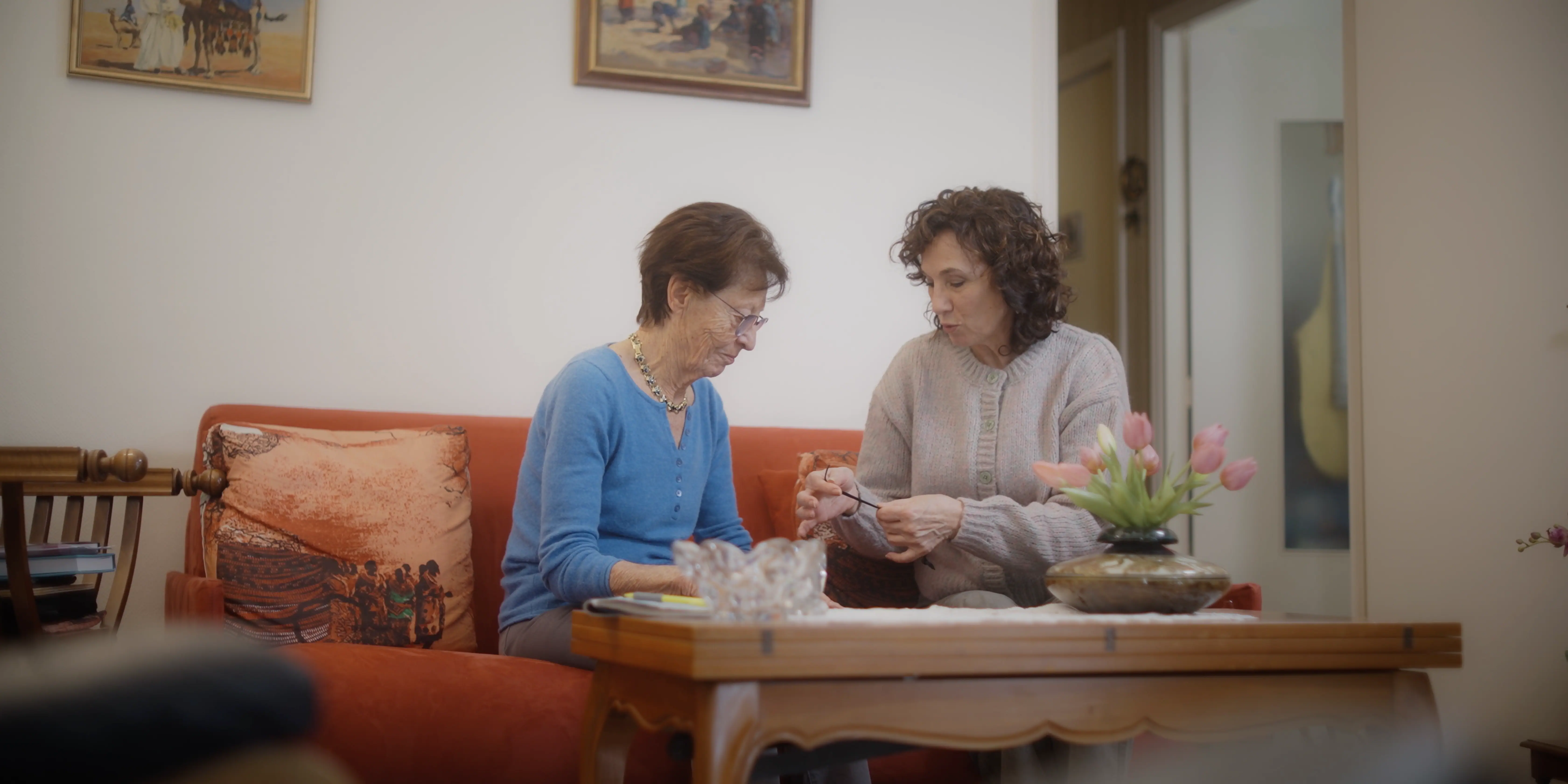 Two women sit on a red sofa in a cozy living room, engaged in conversation, with vibrant artwork on the walls and a vase of pink tulips on the wooden coffee table, creating a warm and inviting atmosphere.