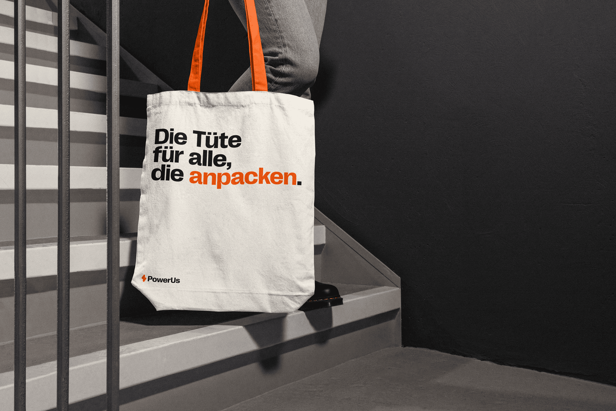 A person walks down a dimly lit staircase while carrying a white tote bag with orange handles, featuring the text "Die Tüte für alle, die anpacken," and the branding of "Powerus" at the bottom.