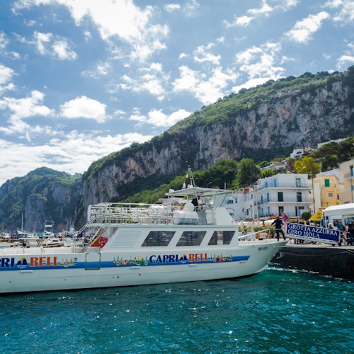 סירה בשם "Capri Bell" עגנה ליד קו חוף עם צוקים, בתים ושלט לסיורי "Grotta Azzurra" תחת שמיים מעוננים חלקית.