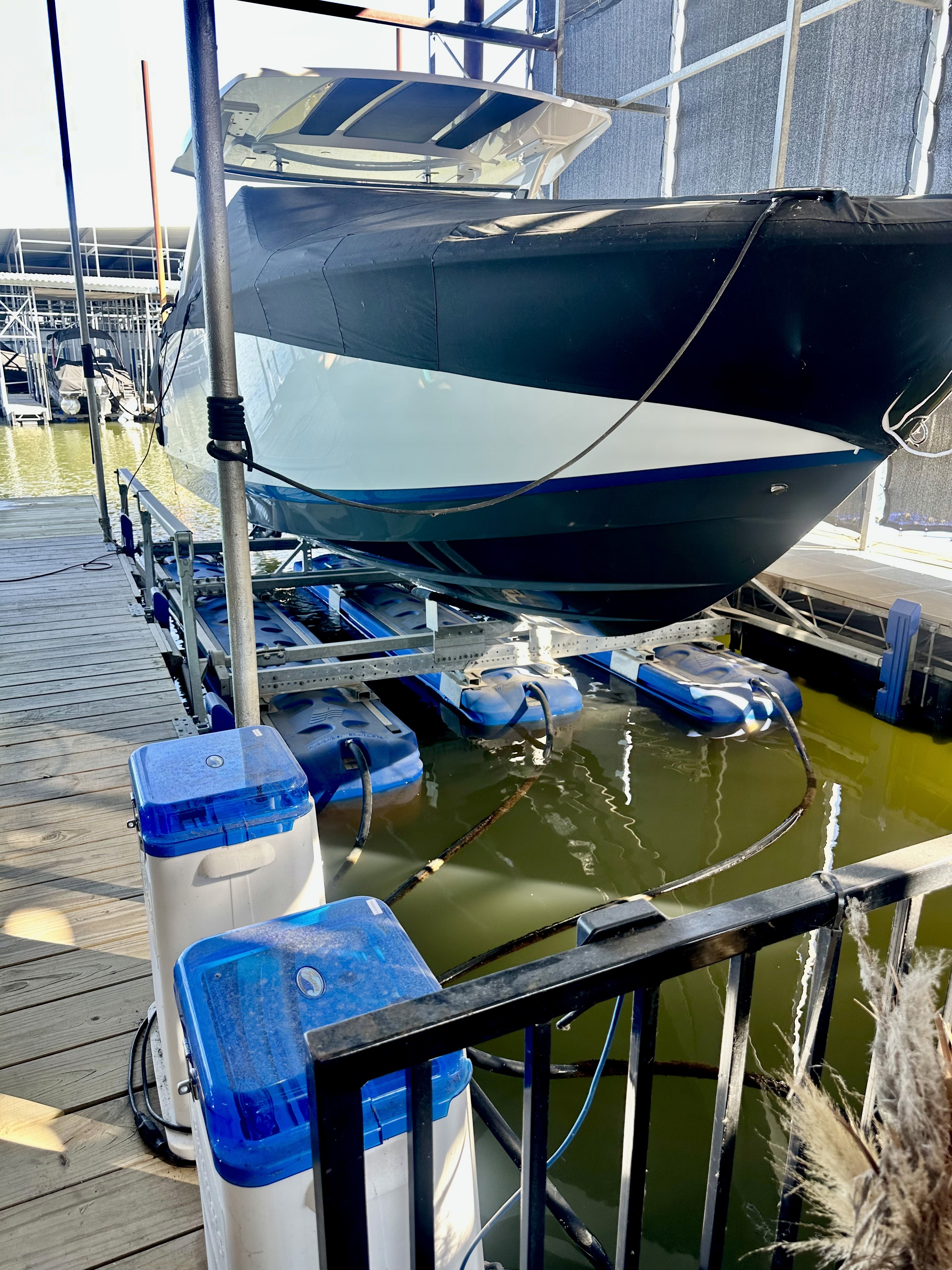 A sleek black and white boat is elevated on a lift in a marina, with surrounding blue and white flotation devices visible, set against a backdrop of docks and parked watercraft.