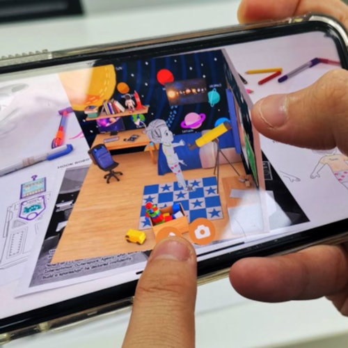 Two hands hold a smartphone displaying an augmented reality room with colorful objects, toys, and a checkered rug.
