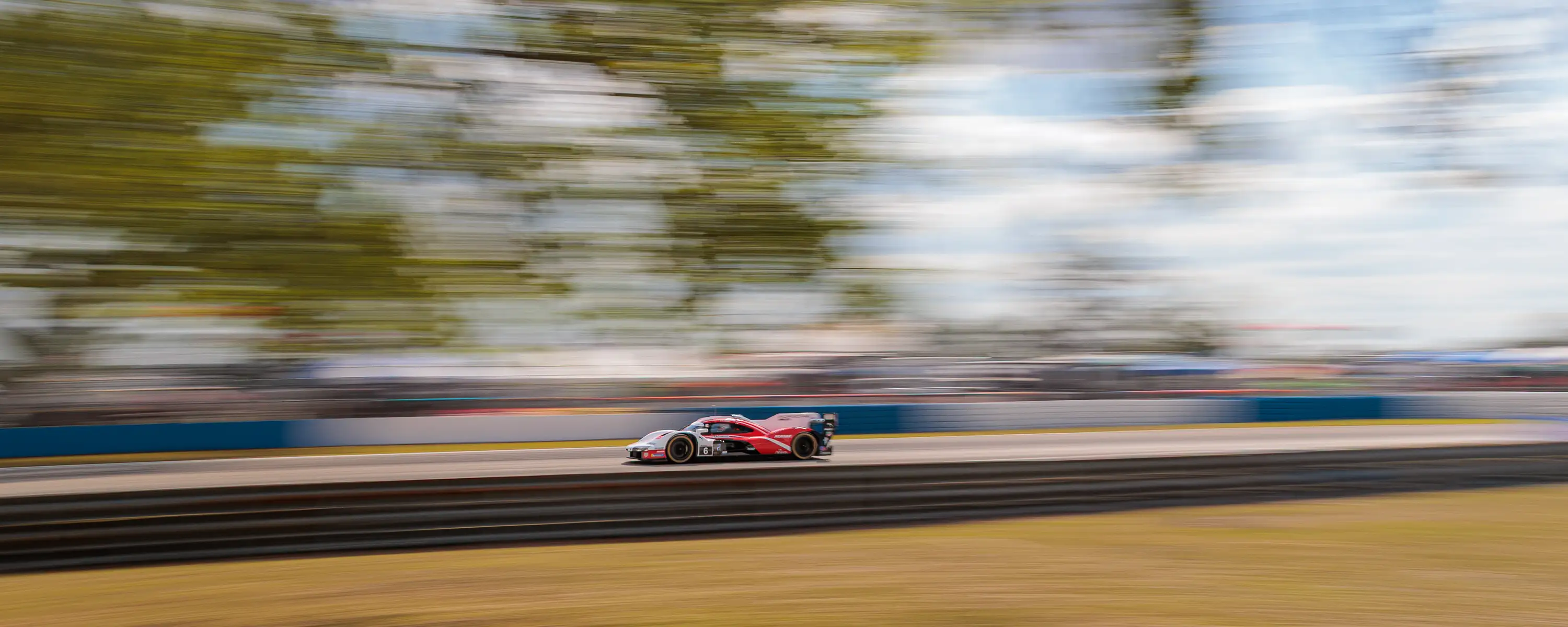 A red Porsche racing car speeds down a track, against a blue sky and trees, with a motion blurred background.