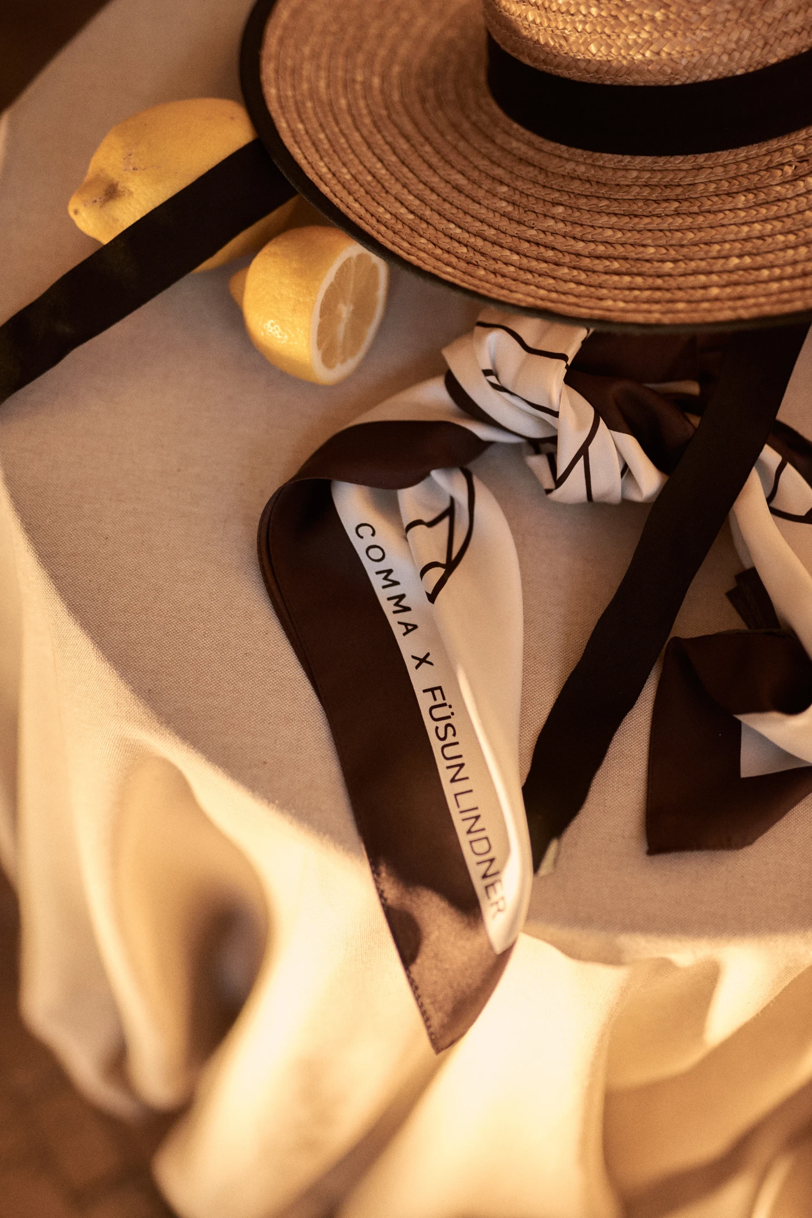 Straw hat, lemons and black and white silk scarves with Comma x Füsun Lindner lettering on a table.