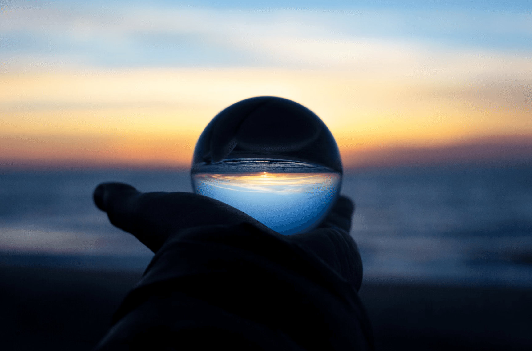 A hand holding a glass ball that reflects a sunset over the sea