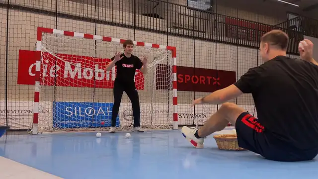Handball goalkeeper facing a seated coach holding a small white ball during a reaction drill in front of the goal.