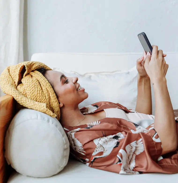 A woman relaxing on a couch at home, smiling while looking at her phone in a calm, unhurried moment.
