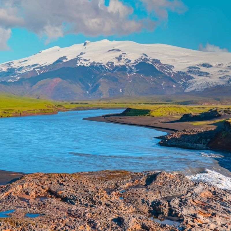 Snow-covered Mýrdalsjökull glacier above green summer slopes, with a blue river winding between dark gravel banks.