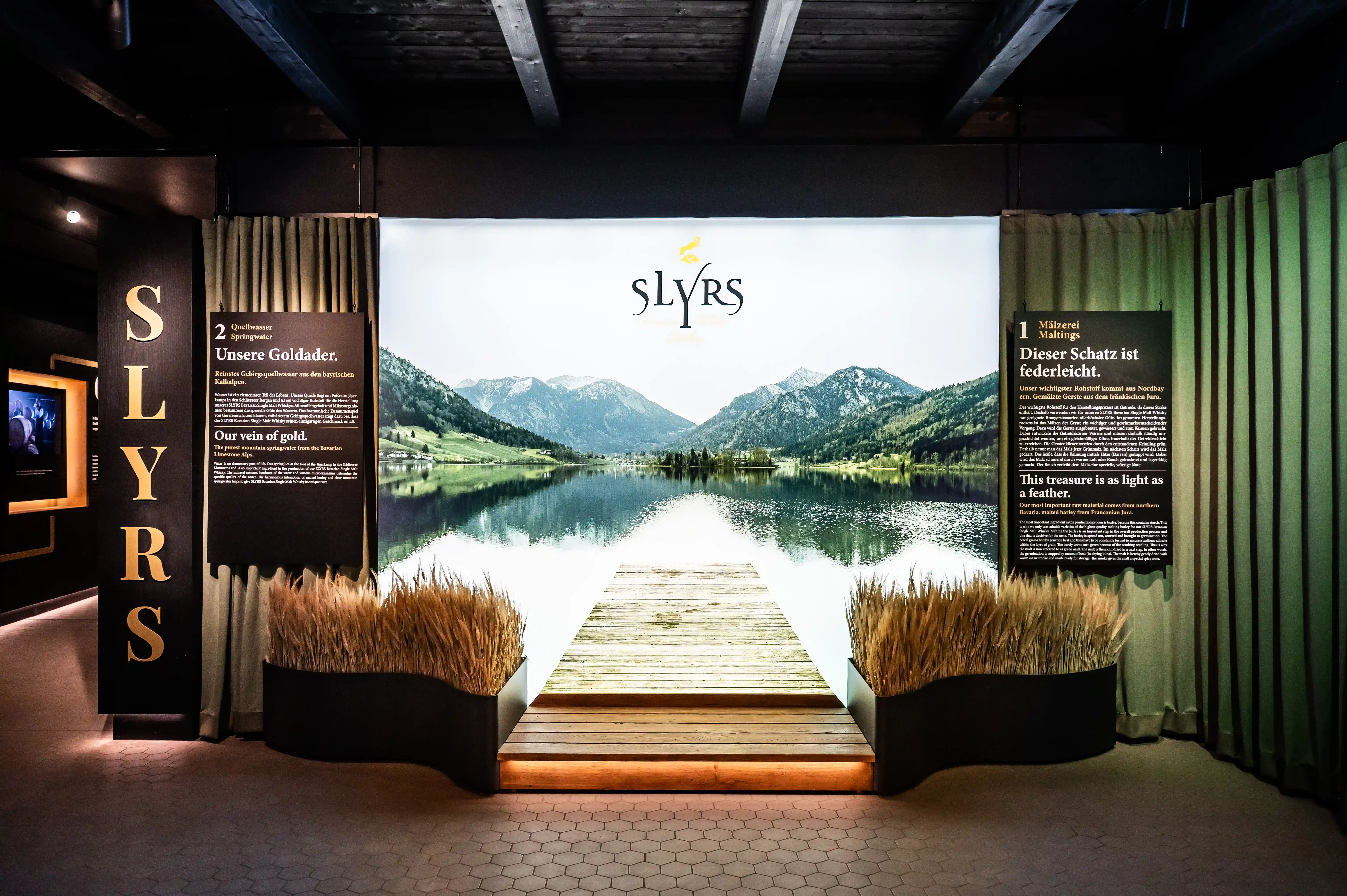 Backlit lake panorama with wooden jetty and SLYRS logo framed by golden lettering pillars and grasses at the entrance.