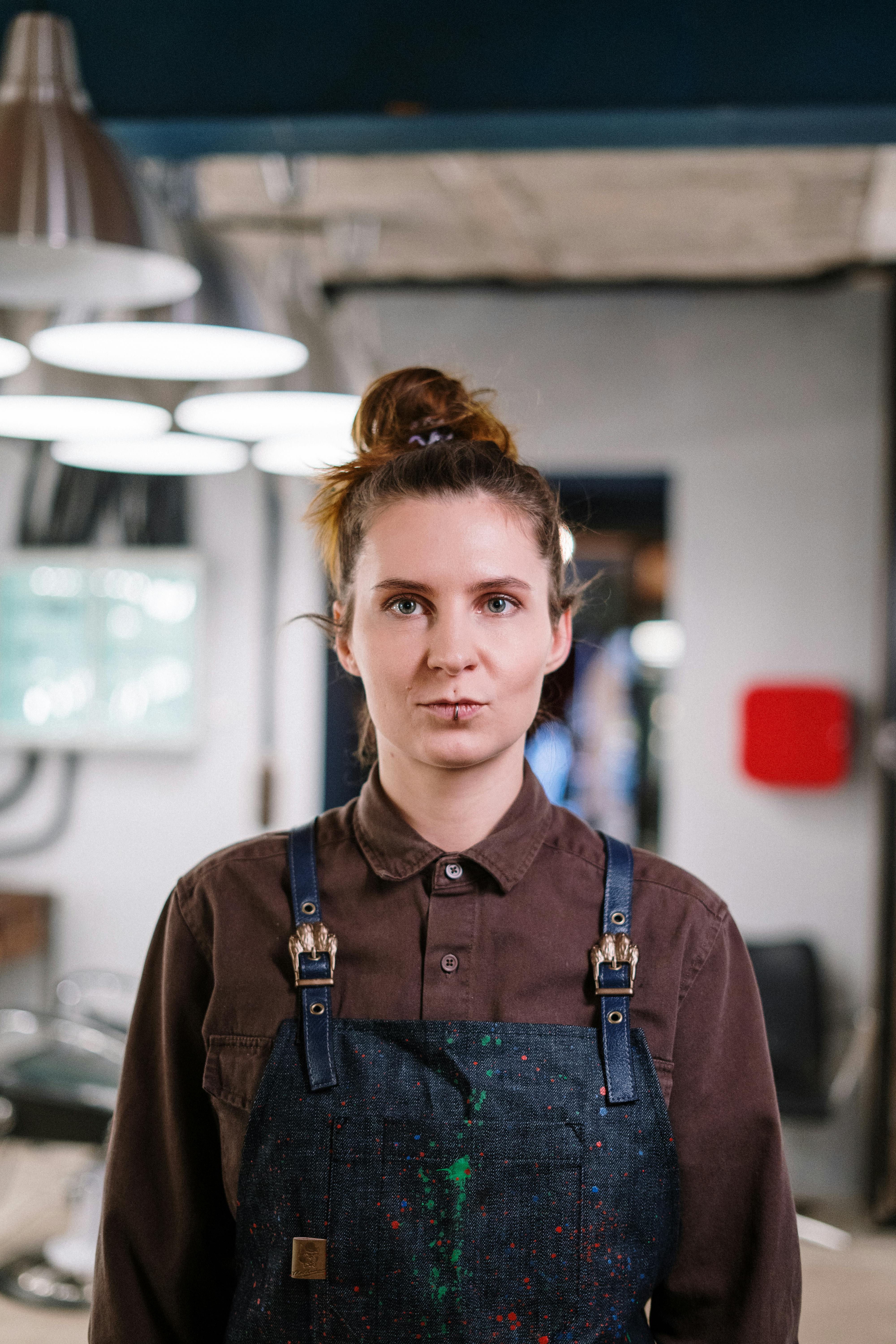 A woman with a lip piercing looking directly at the camera wearing a brown shirt and blue apron.