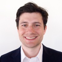 A person with a friendly smile is wearing a black blazer and white shirt, set against a plain white background, exuding a professional and approachable demeanor.