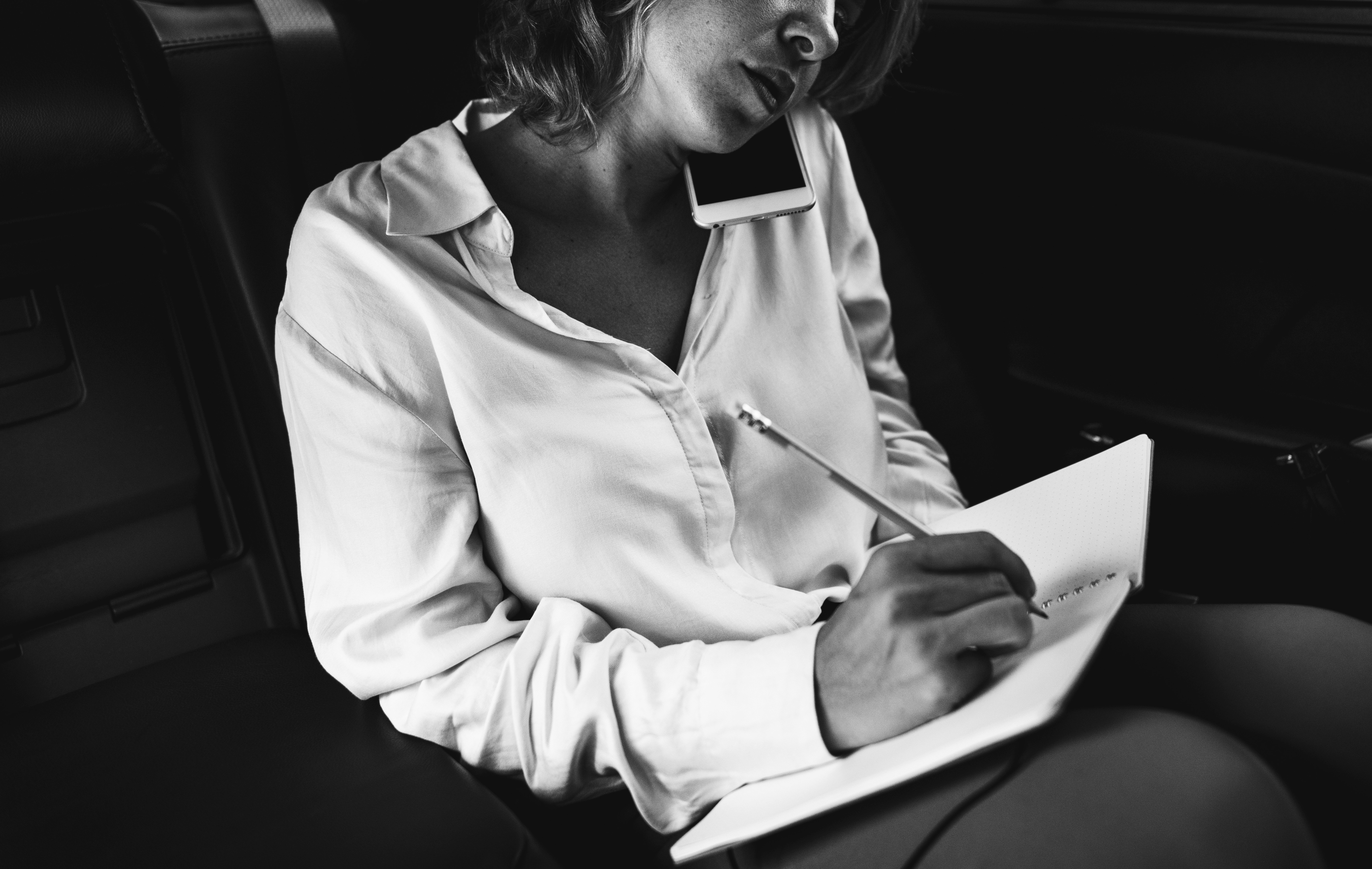 A seated person wearing a shirt in a light shade of grey, writing on a tablet or notebook, in a black-and-white photo.