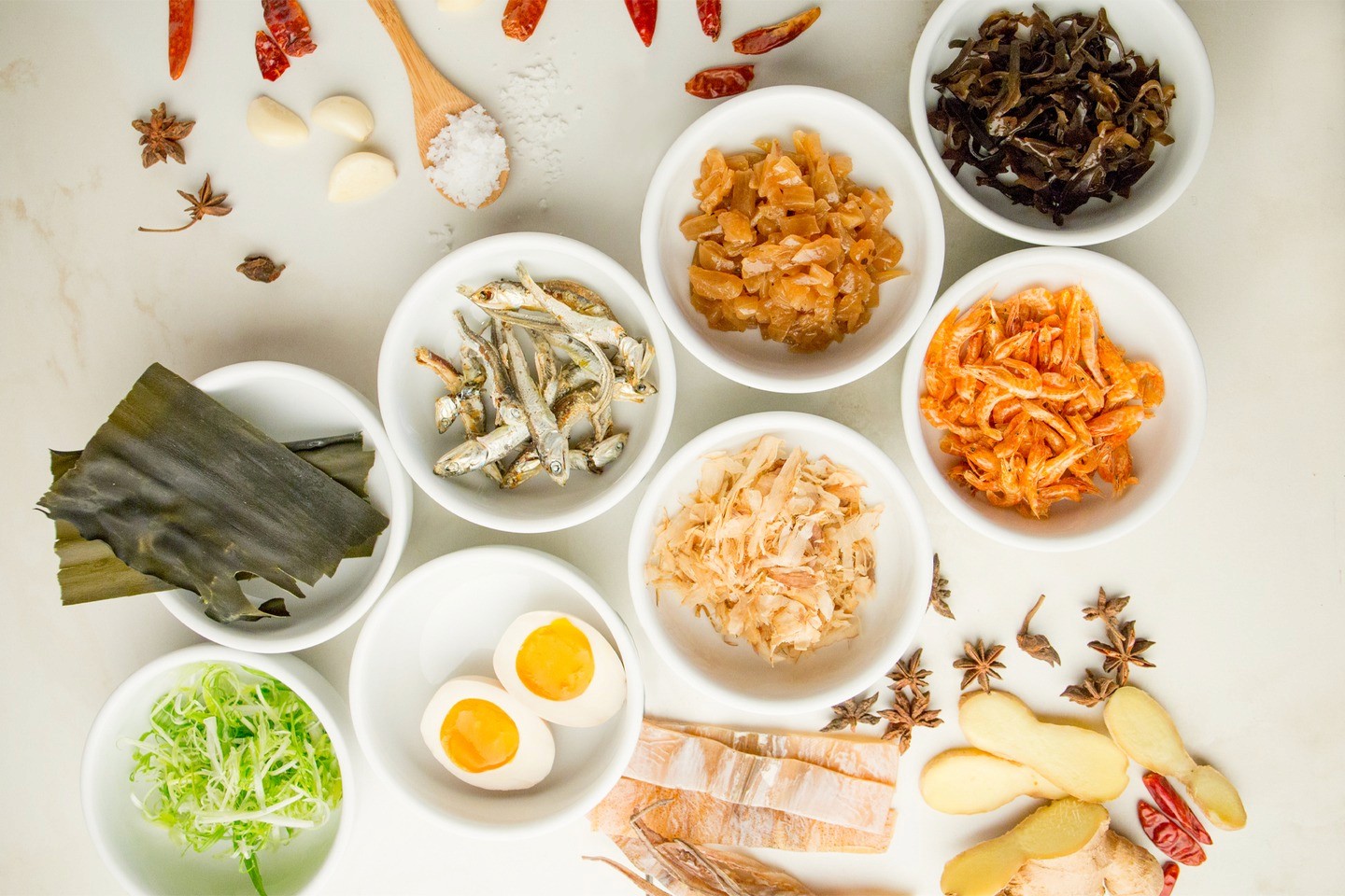 Assorted ramen ingredients in white bowls including soft-boiled eggs, dried anchovies, kimchi, mushrooms, and spices on a white surface