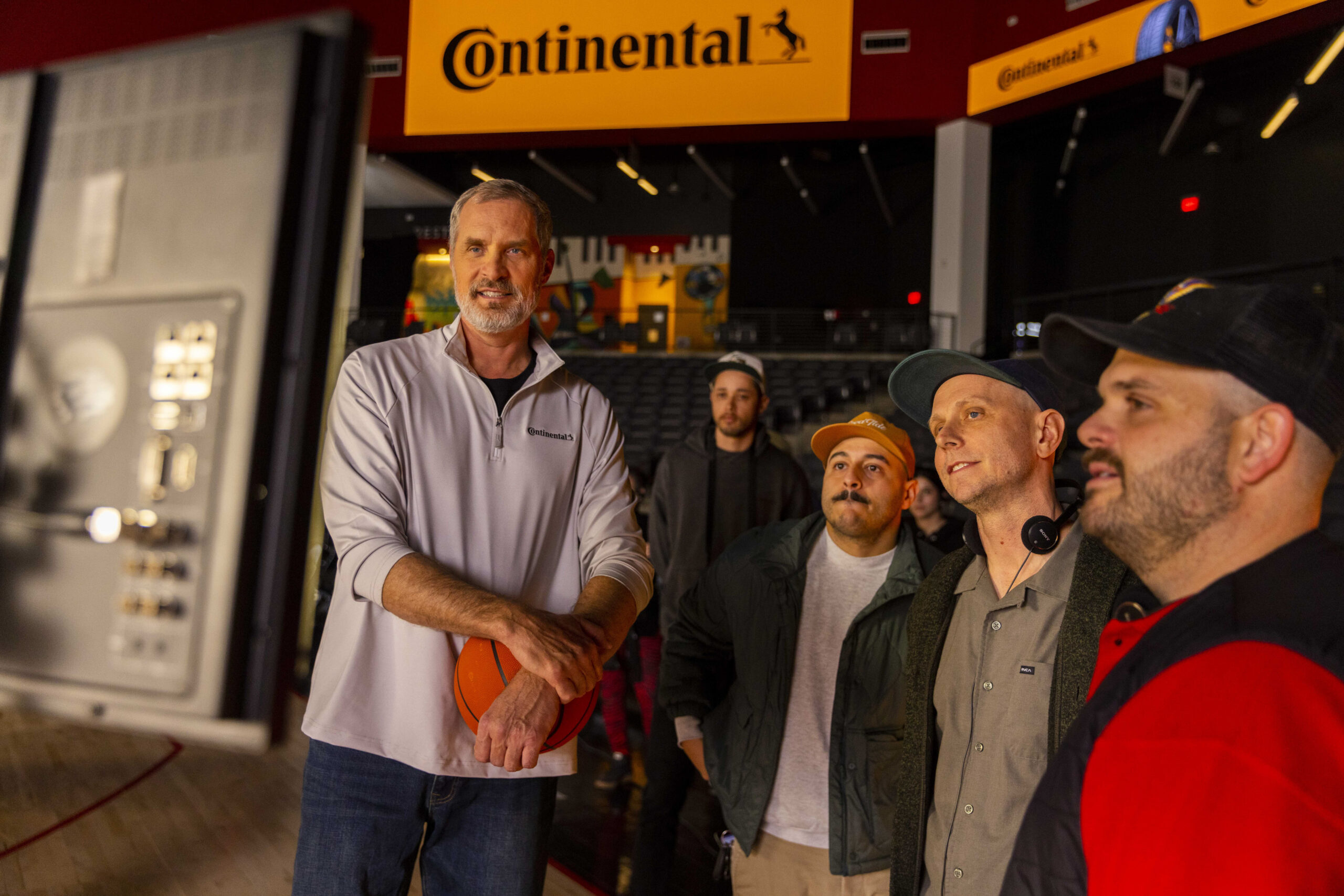 A group of five men stand indoors near a basketball court, with one holding a basketball. Bright Continental signage is visible in the background. They appear to be attentively watching something off camera.