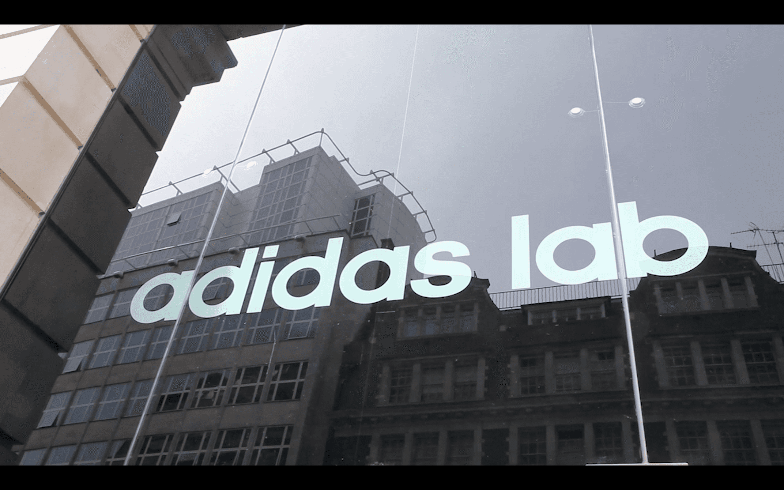 The image shows a modern urban street scene reflected in the large, glass windows of a building with "adidas lab" written prominently in bold white letters, capturing a blend of city architecture and brand presence.