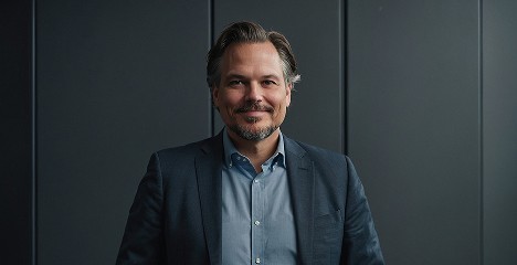 A man in a dark suit and light blue shirt stands against a dimly lit, modern gray panel backdrop, exuding confidence and professionalism.