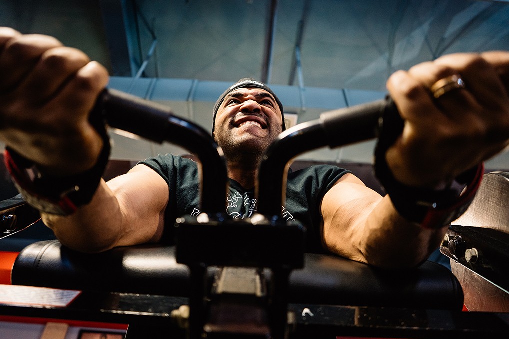 A man is seated on a black weight machine, gripping the handles with both hands. He wears a dark green t-shirt and a dark-colored baseball cap. His face is visible, showing a broad smile. The machine has black padded handles and a black frame. The background is blurred, showing a blue-gray wall.