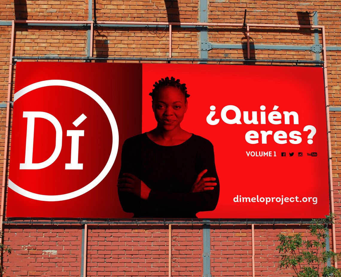 A red billboard displays a woman with dark skin and braided hair, wearing a black long-sleeved top, with her arms crossed. Above her, the white text "¡Quién eres?" is prominent. Below her, the text "VOLUME I" appears on the left, and "dimeloproject.org" is on the right. The billboard is mounted on a red brick wall with visible pipes and supports.