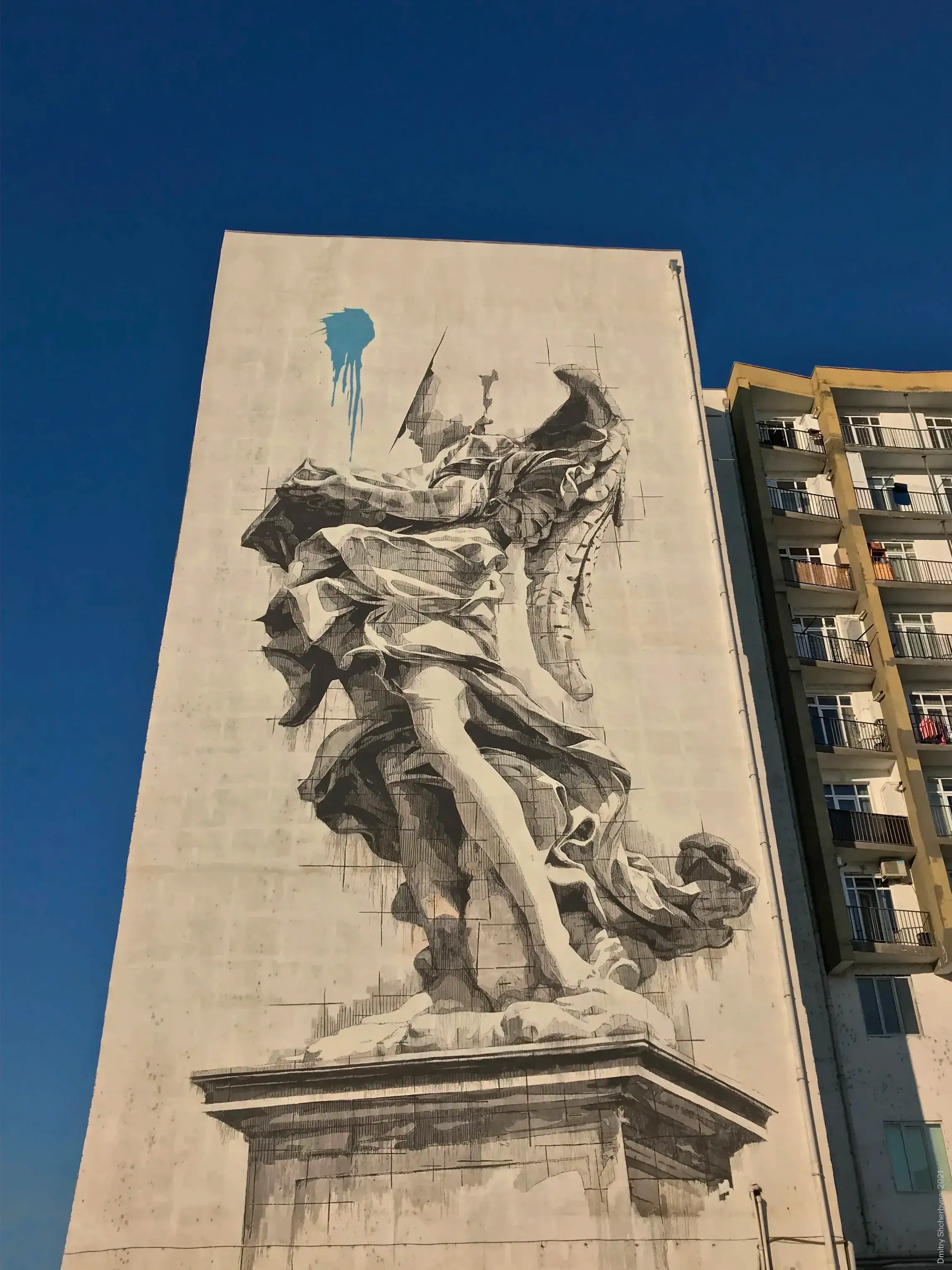 A monochrome black-and-white mural spanning the full height of a building, depicting a Baroque-style winged angel statue on a pedestal in dramatic flowing robes, rendered with fine cross-hatching.