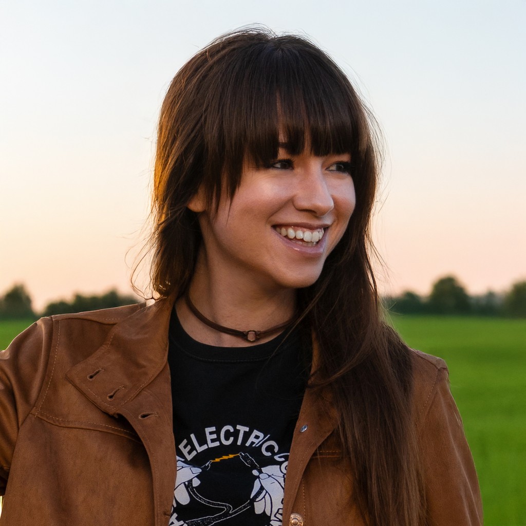Holly Stewart smiling outdoors at sunset, wearing a brown leather jacket and black graphic tee with long dark hair and bangs, standing in front of a green field and soft pastel sky.