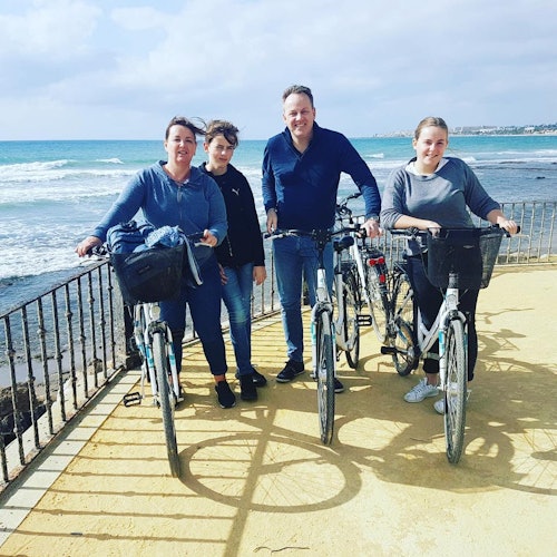 Four people standing with bicycles on a seaside promenade, with the ocean and a partly cloudy sky in the background.