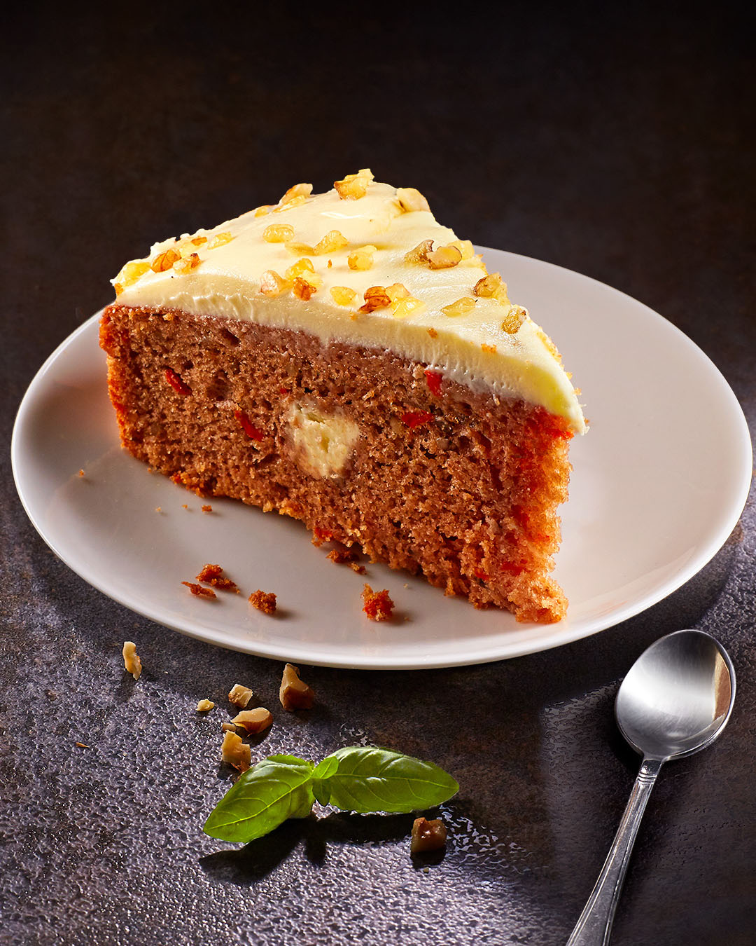 A slice of moist carrot cake with cream cheese frosting, sprinkled with chopped walnuts, is presented on a white plate next to a spoon, complemented by a small mint leaf for garnish.