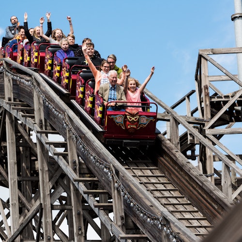 People riding a wooden roller coaster with their arms raised, expressing excitement and joy, under a clear blue sky.