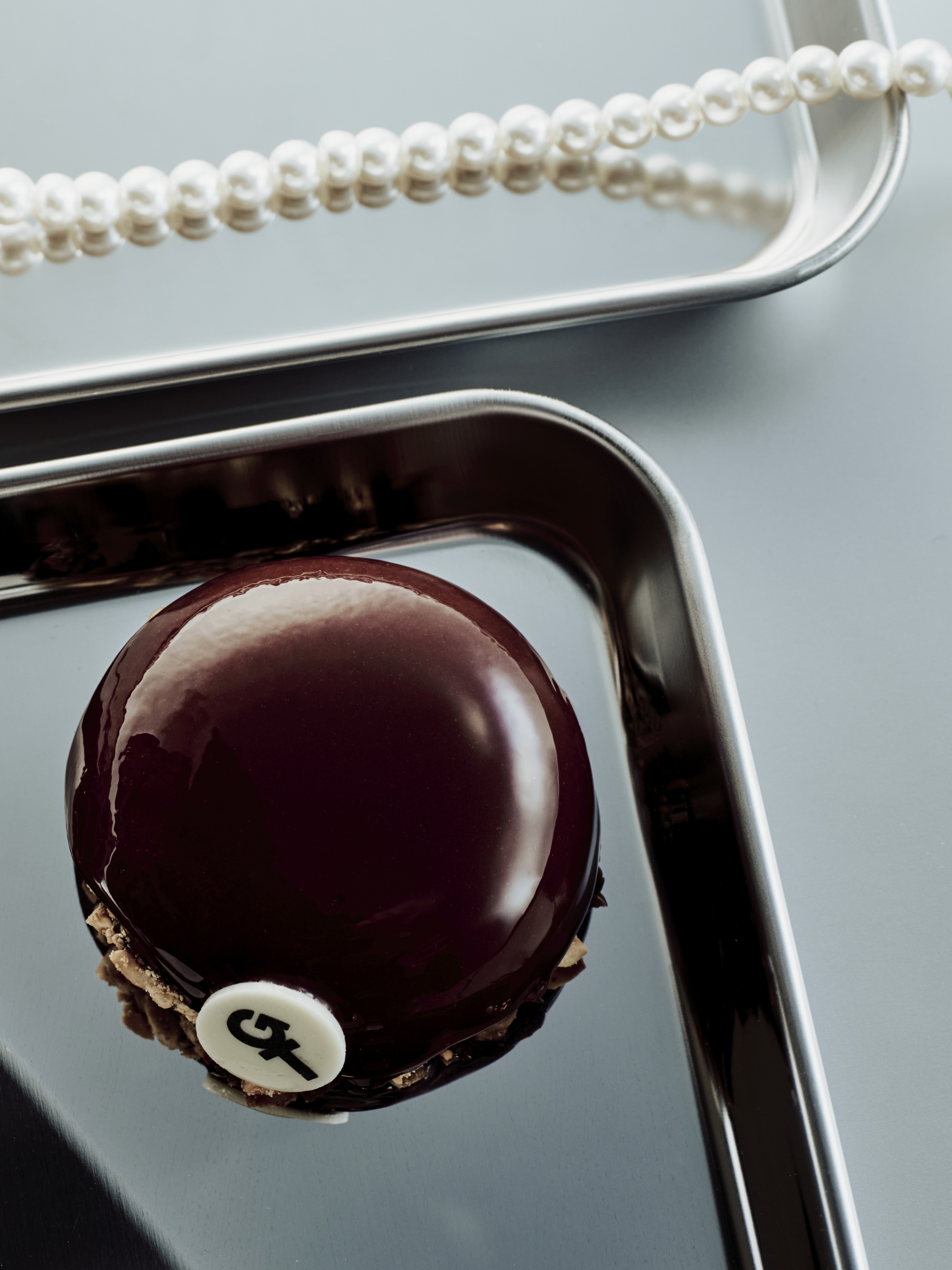 Dôme glacé au chocolat lustré, orné d’un logo 'GT', posé sur plateau métallique avec perles en arrière-plan