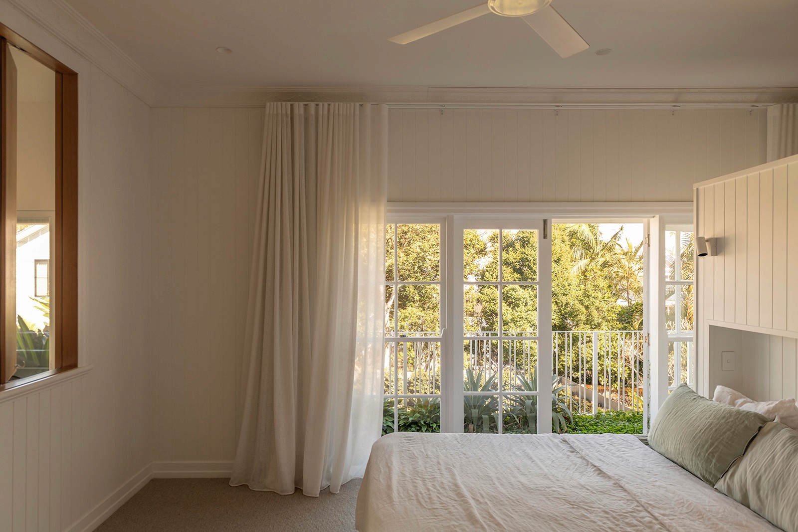 Light-filled bedroom with soft white curtains, timber details, and French doors opening to leafy balcony views.