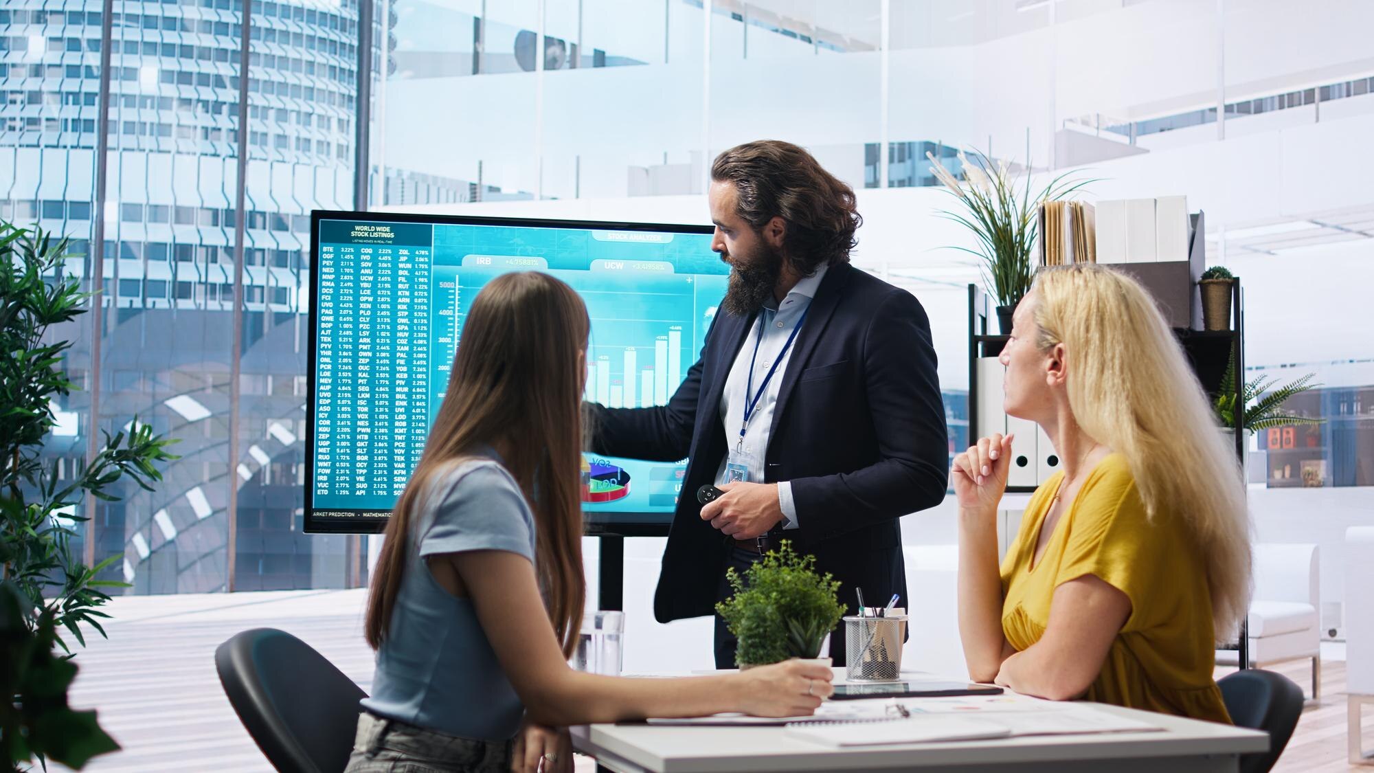 Someone stands before a large display, presenting charts, while two others sit at a table listening. 